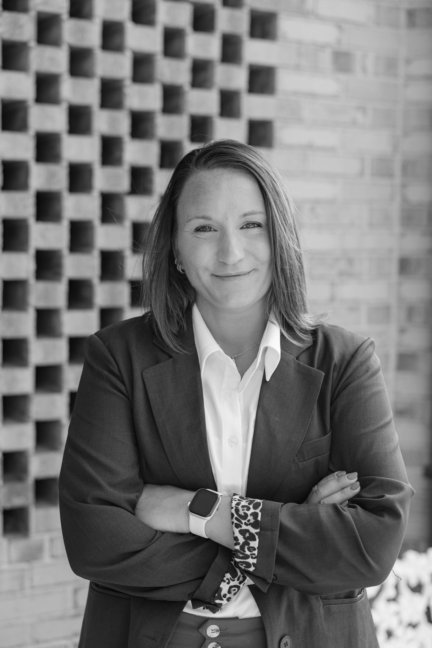 Black and white professional headshot of a woman in a blazer with arms crossed, smiling confidently outdoors.