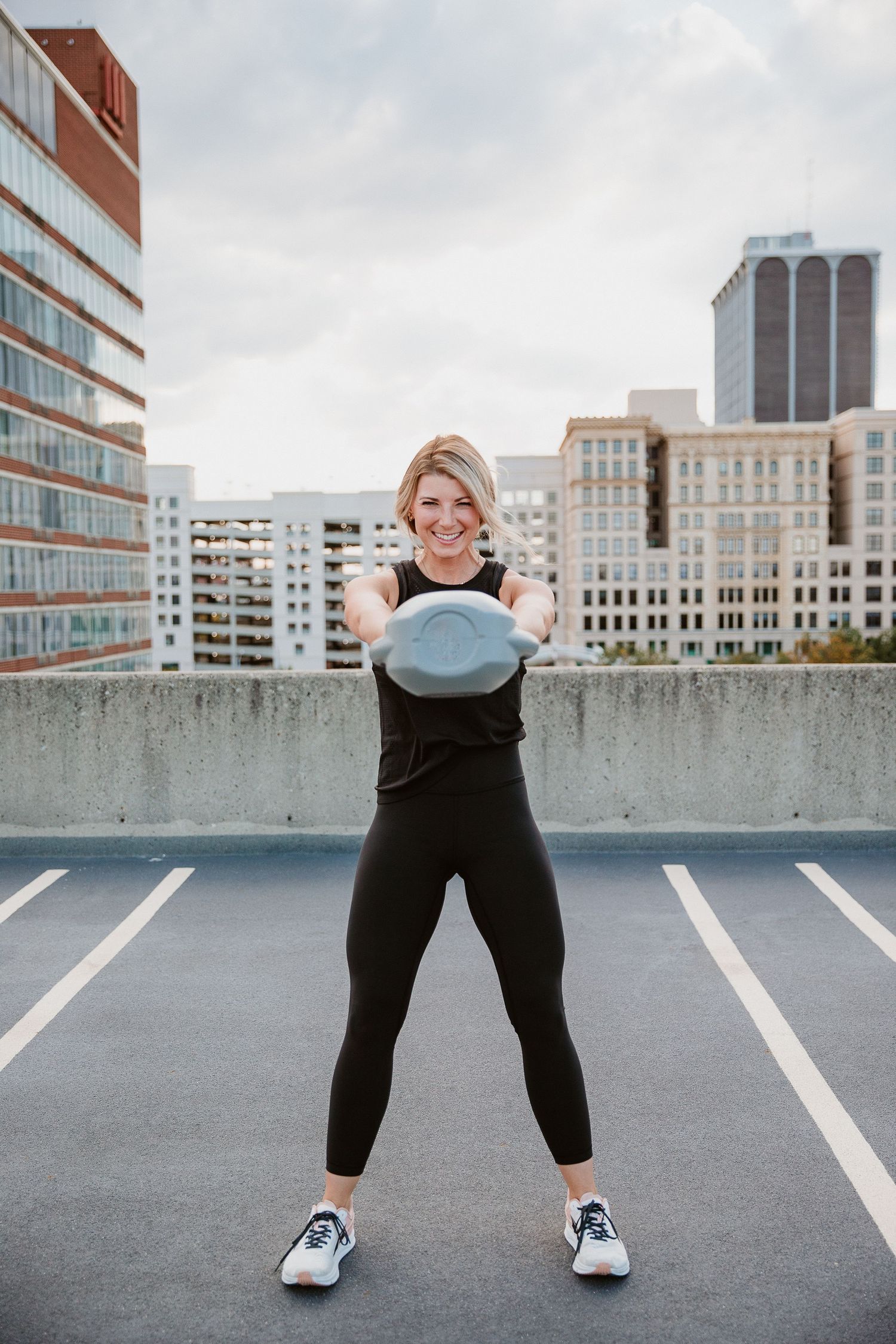 Woman in black athletic wear doing kettlebell swings on a rooftop parking lot with city buildings in the background. Katie Chamberlain
