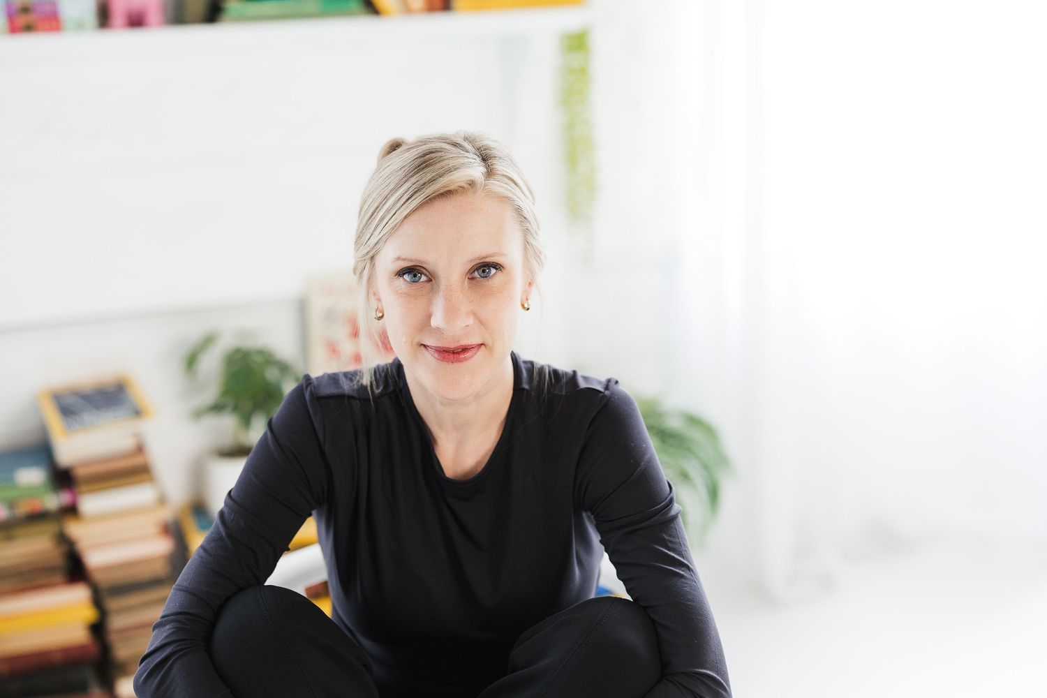 Blonde woman in black outfit sitting cross-legged in a bright room with bookshelves in the background. Katie Chamberlain Photography