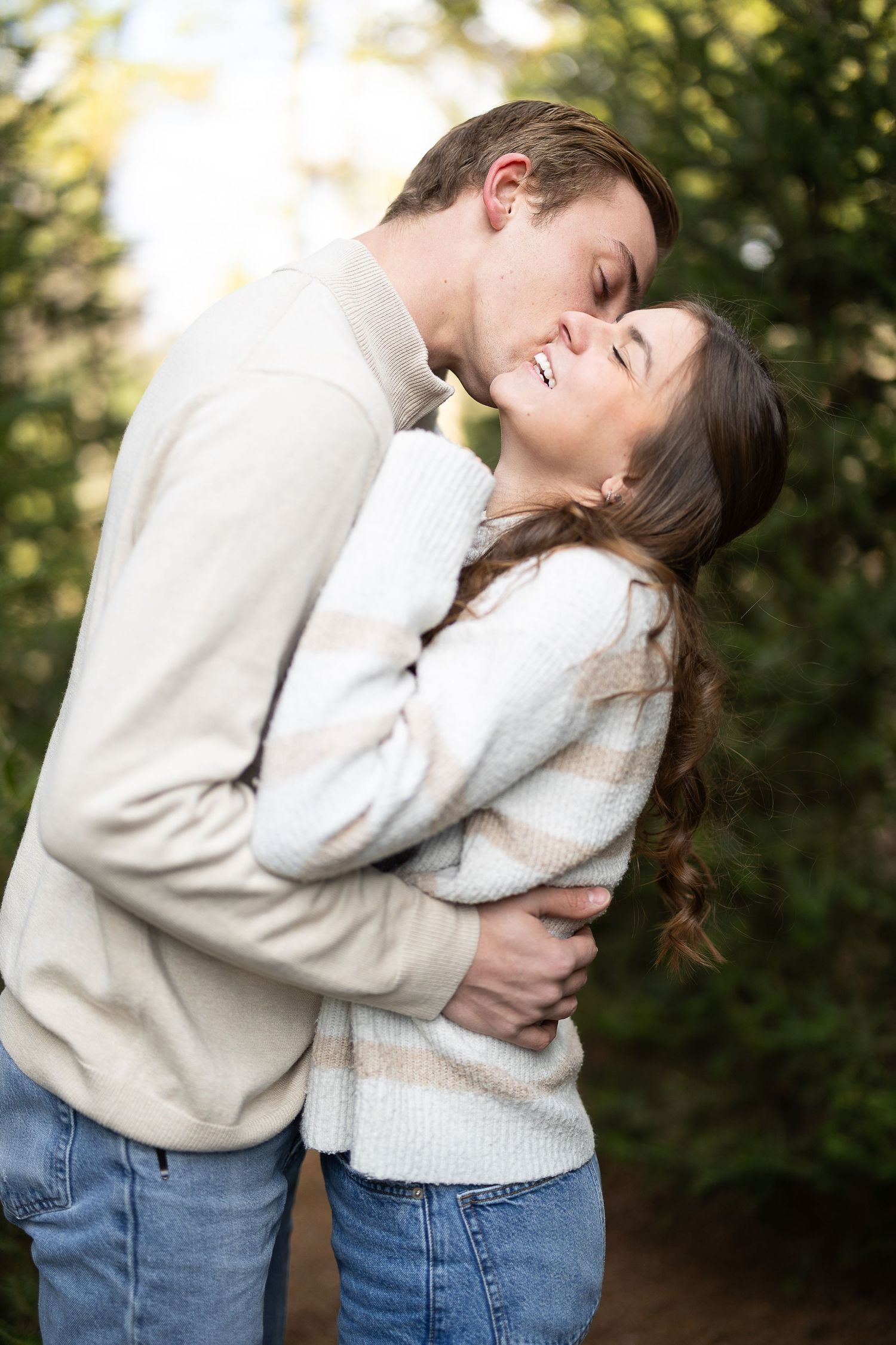 A couple embracing outdoors, man kissing woman's cheek, both wearing cream sweaters and jeans with green trees behind. Katie Chamberlain
