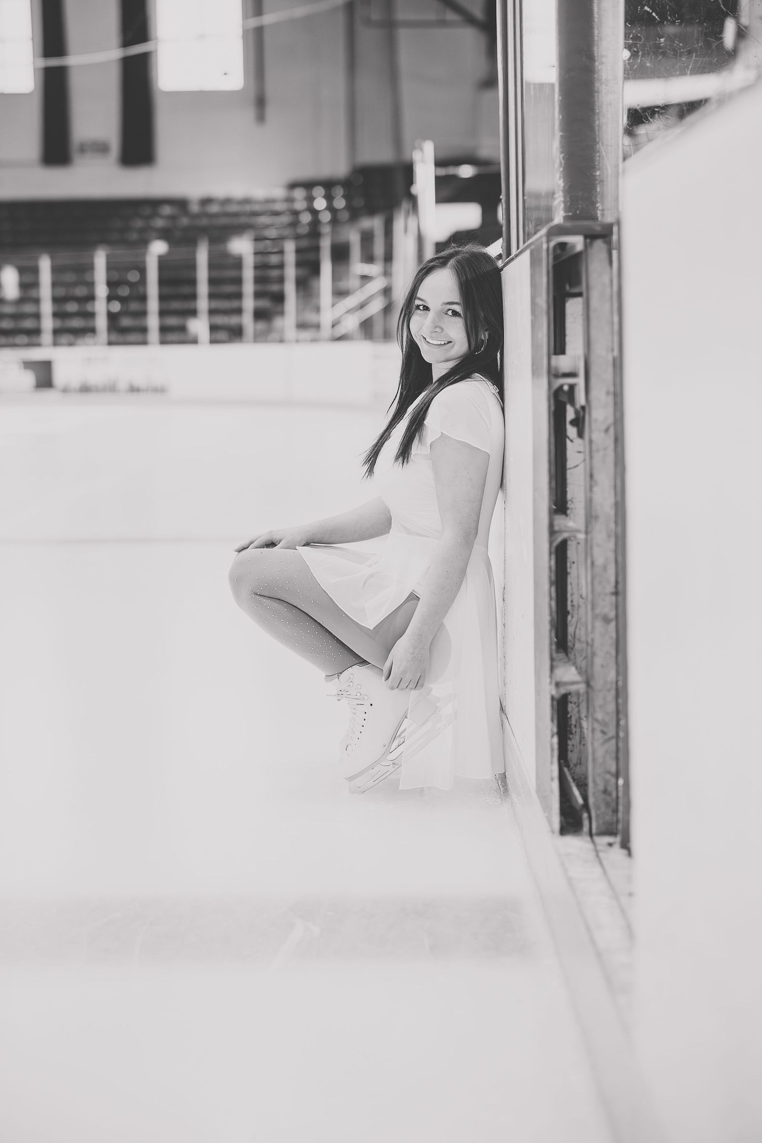 Young woman in white dress and ice skates sitting rinkside at an indoor ice skating rink, black and white photo. Katie Chamberlain Photograp