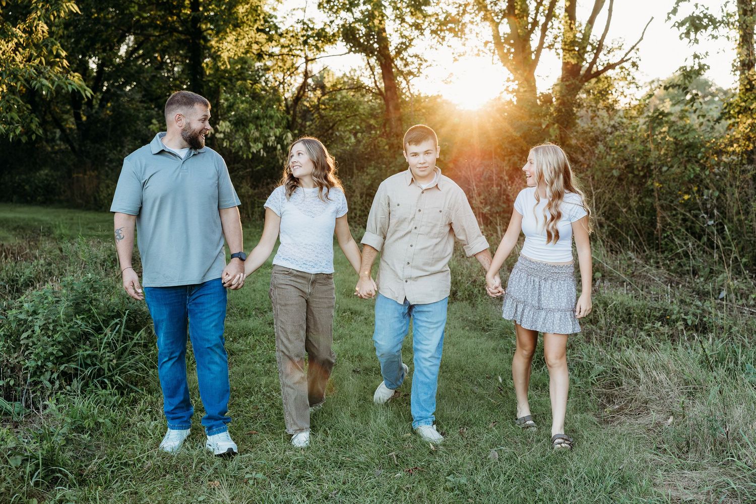 Family of four holding hands walking through a sunlit field with golden hour light filtering through trees.