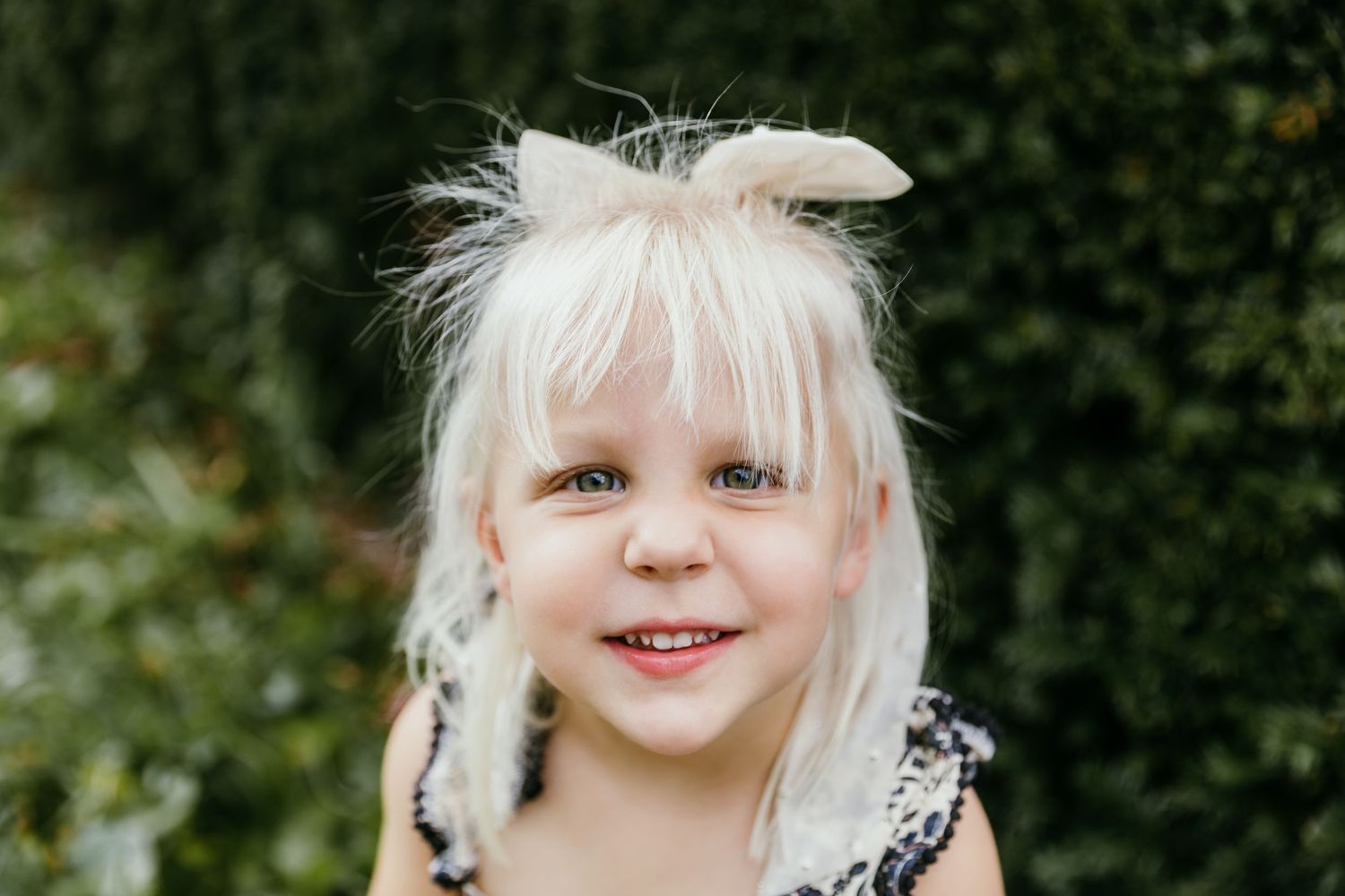 Smiling young girl with blonde hair and a bow, posing outdoors in front of green bushes. Katie Chamberlain Photography
