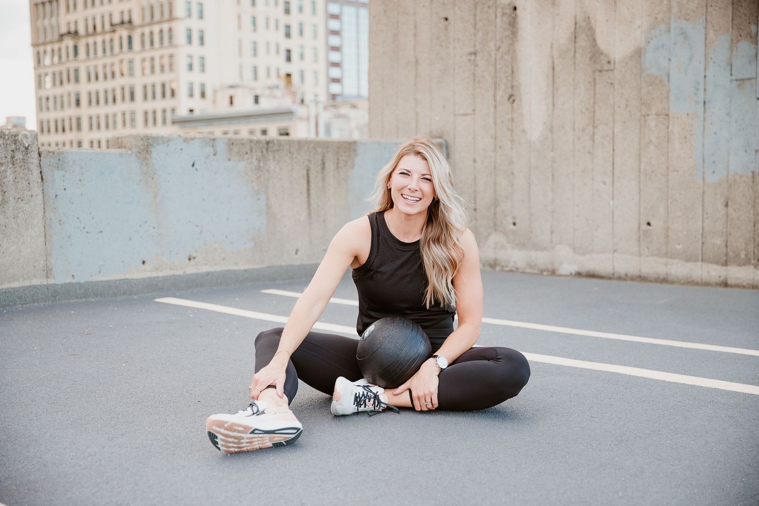 Smiling blonde woman in black athletic wear sitting on rooftop parking lot with city buildings in background. Photo by Katie Chamberlain