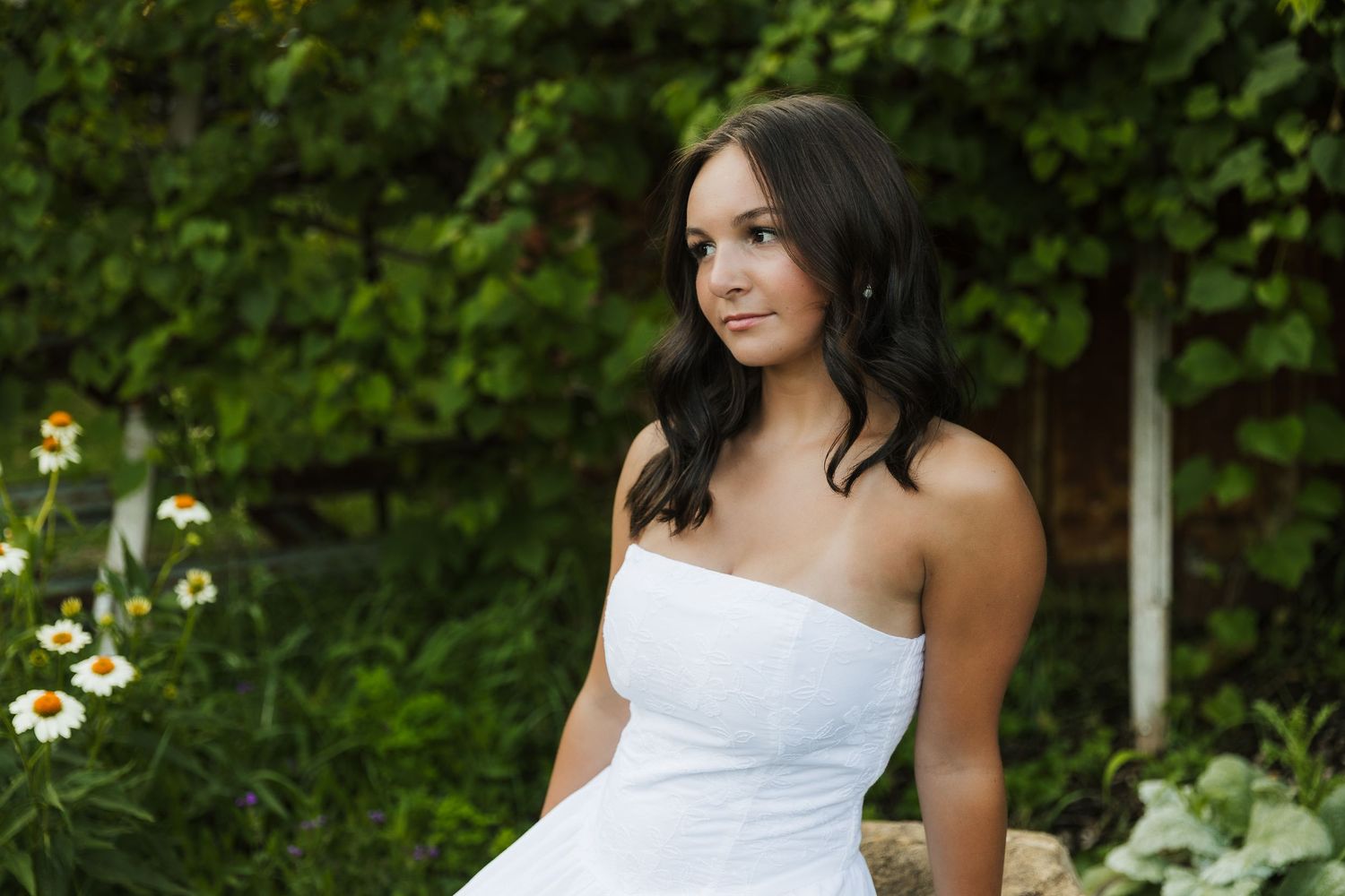 Young woman in white strapless dress posing outdoors among green ivy and white wildflowers. Katie Chamberlain Photography