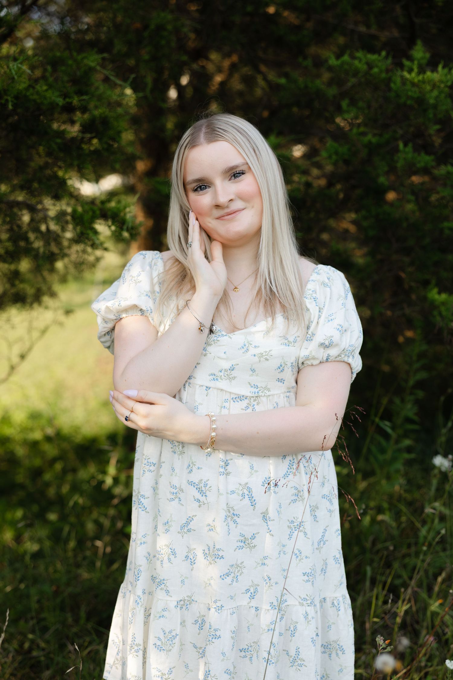 Young blonde woman in a floral white dress poses outdoors among green trees in a serene garden setting.
