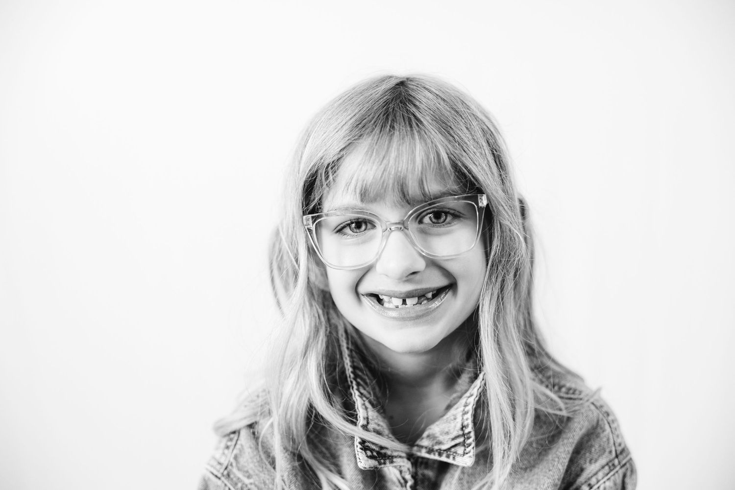 Black and white portrait of a smiling young girl with long blonde hair and glasses, wearing a denim jacket.
