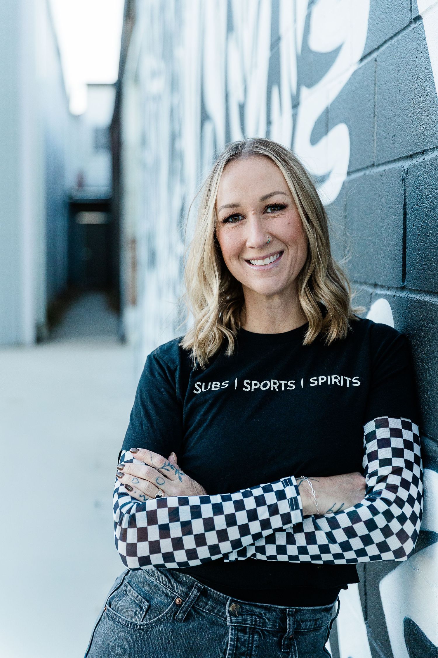 Smiling blonde woman in black Subs Sports Spirits t-shirt with checkered sleeves, arms crossed, posing in urban alley. Katie Chamberlain