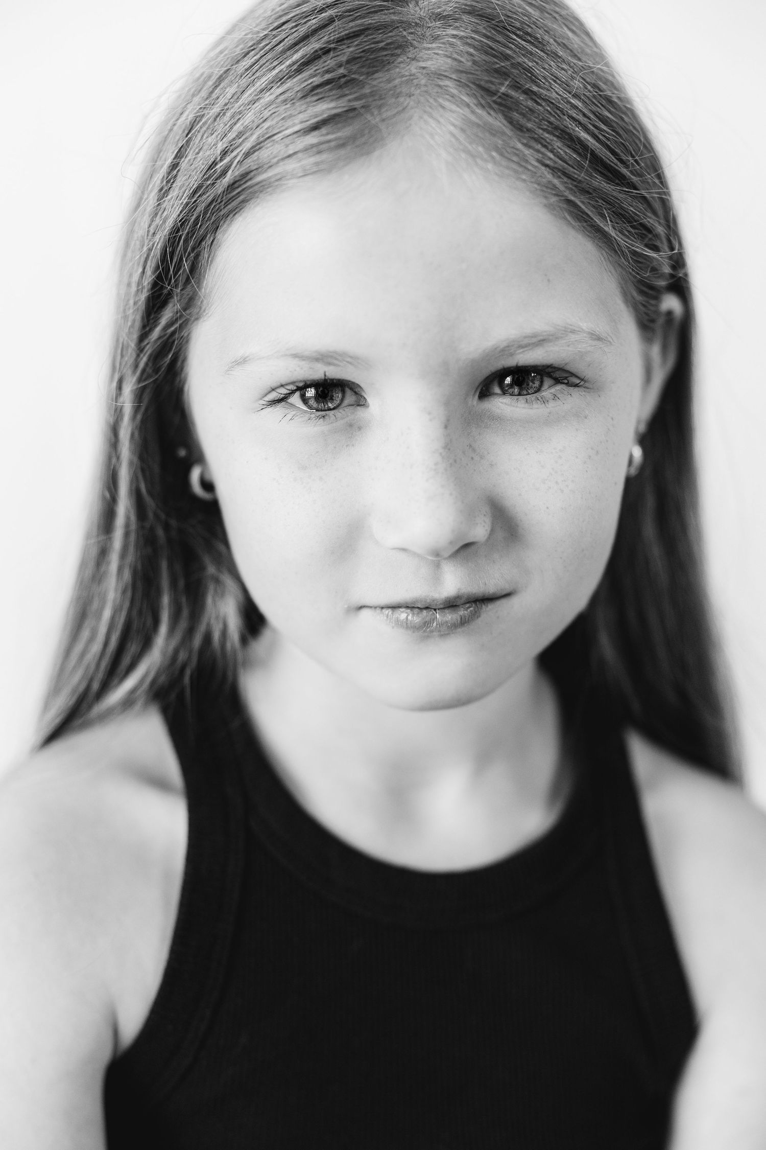 Black and white portrait of a young girl with long hair, freckles, and small earrings wearing a black sleeveless top.