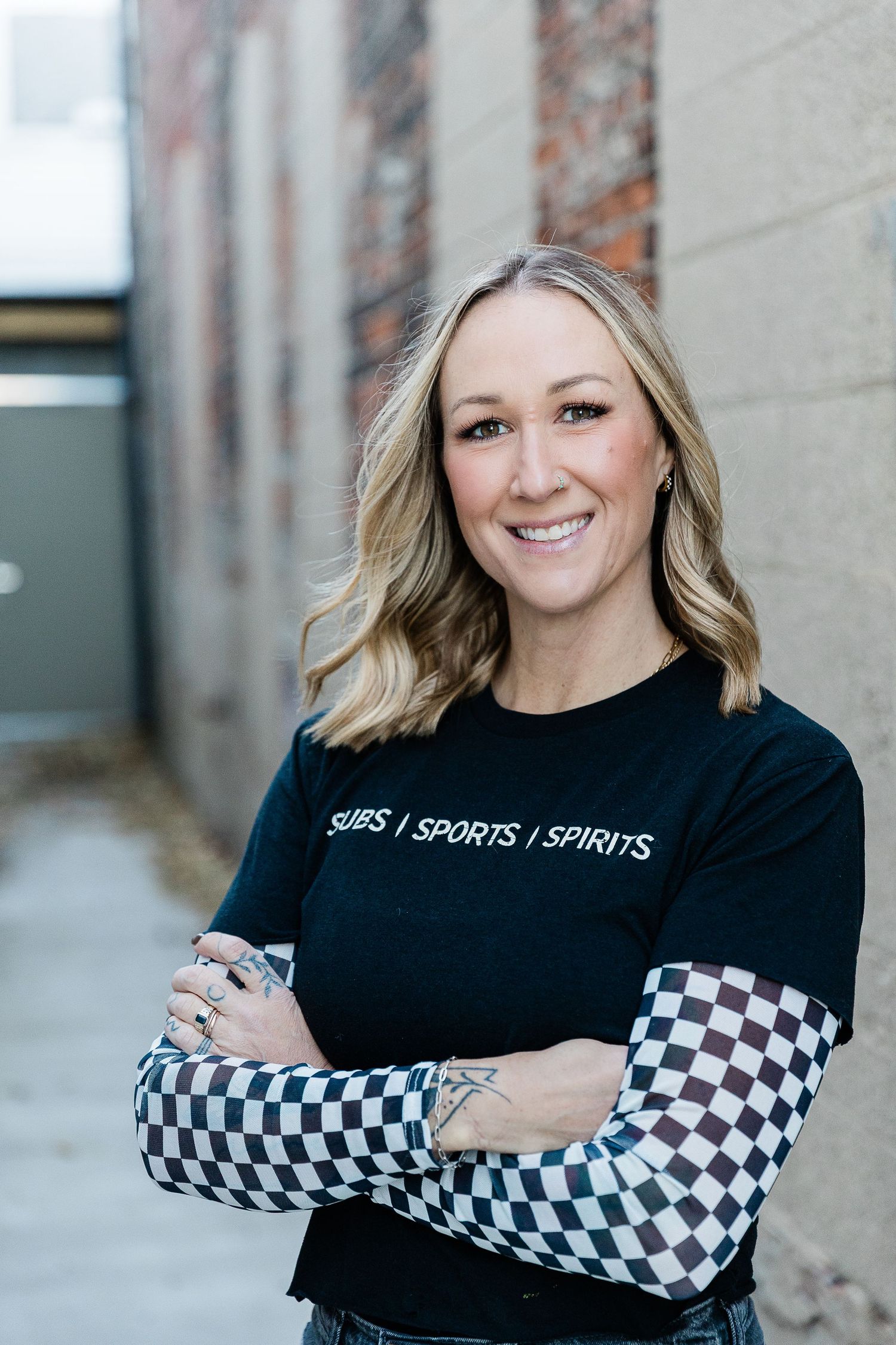 Smiling blonde woman in black Subs Sports Spirits t-shirt with checkered sleeves, arms crossed, posing in urban alley. Katie Chamberlain