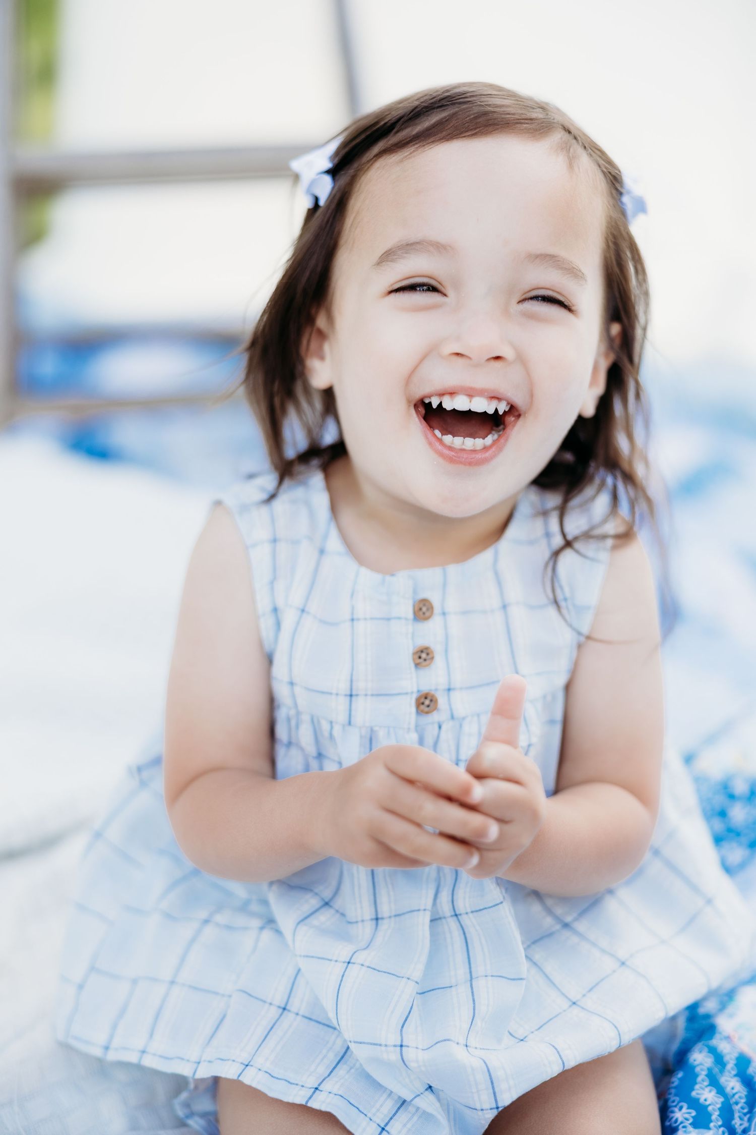 A joyful toddler girl in a blue plaid dress laughs brightly, wearing blue hair bows outdoors.