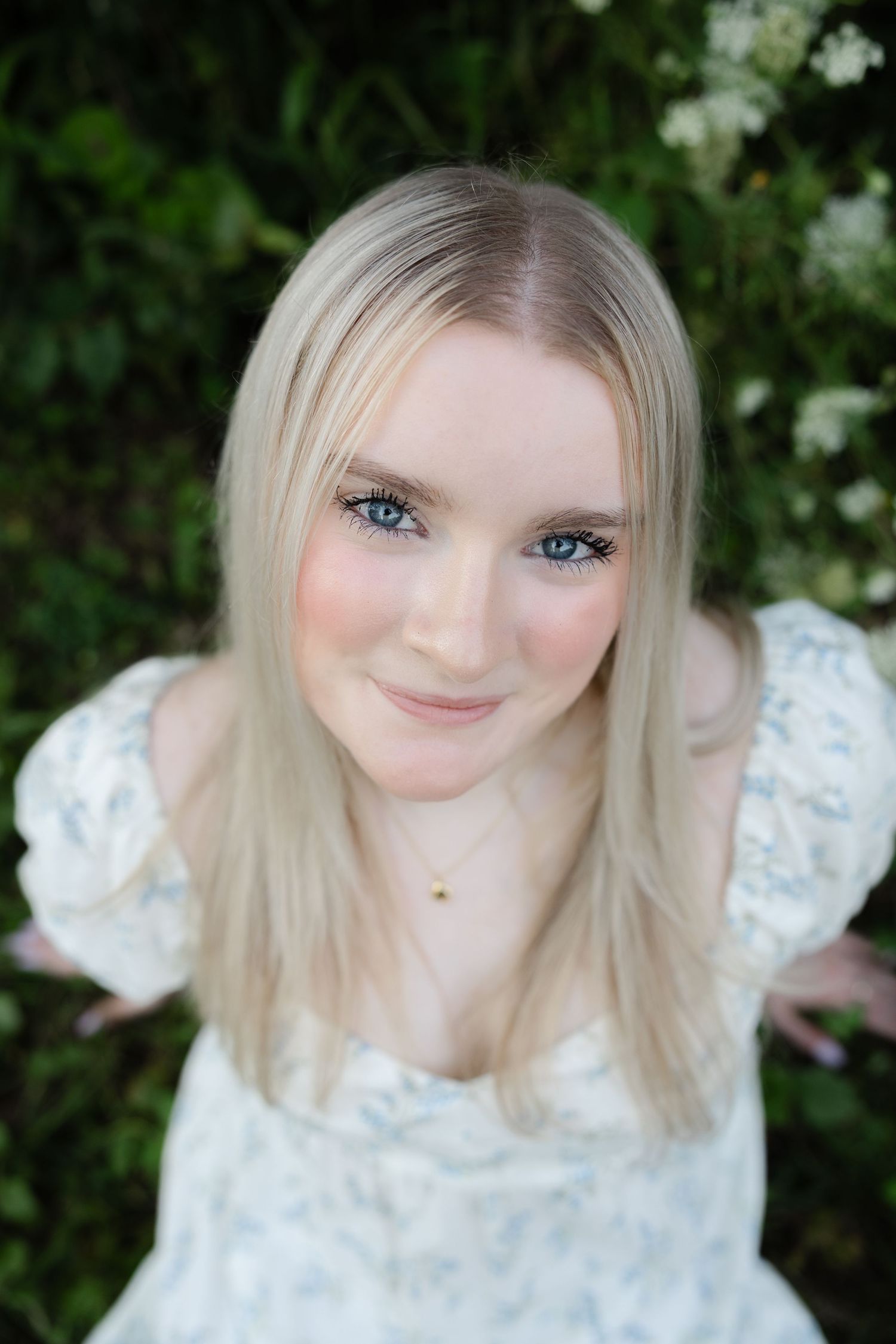 Young blonde woman with blue eyes wearing a white floral dress, smiling outdoors with green foliage background.