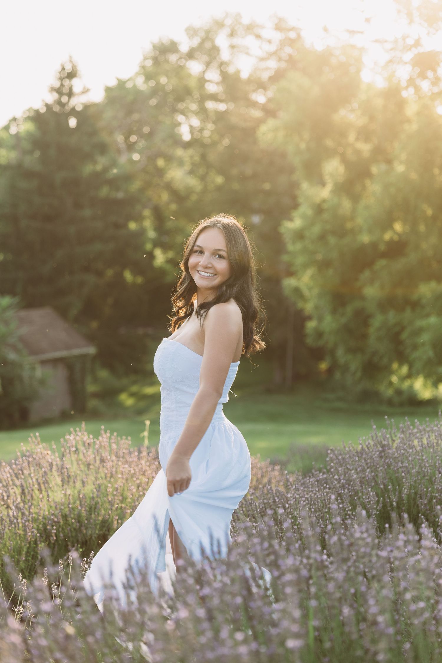 Young woman in white dress smiling in lavender field at golden hour sunset with green trees background. Katie Chamberlain Photography