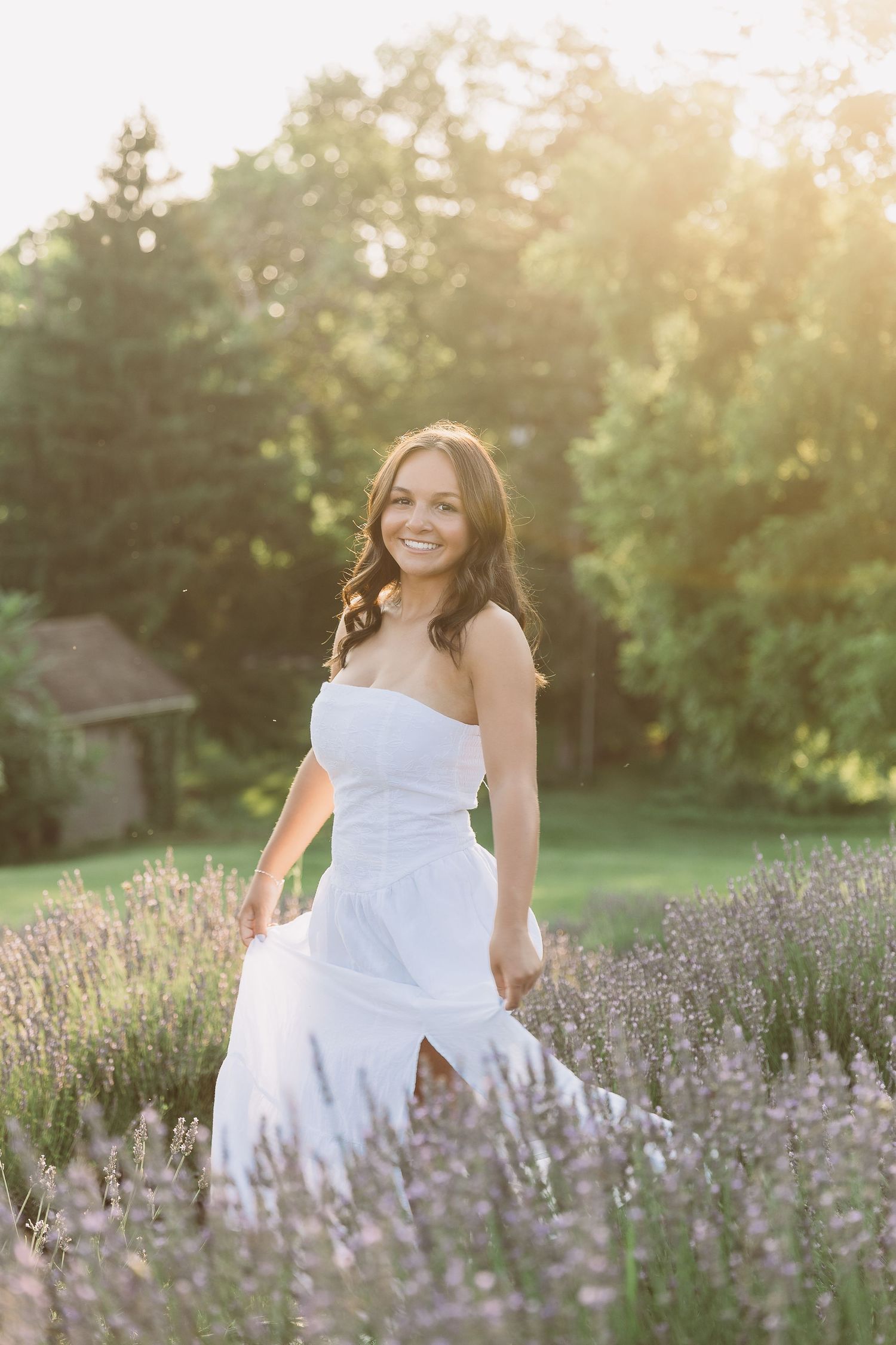 Smiling bride in white strapless gown standing in lavender field with golden sunlight and green trees in background. Katie Chamberlain