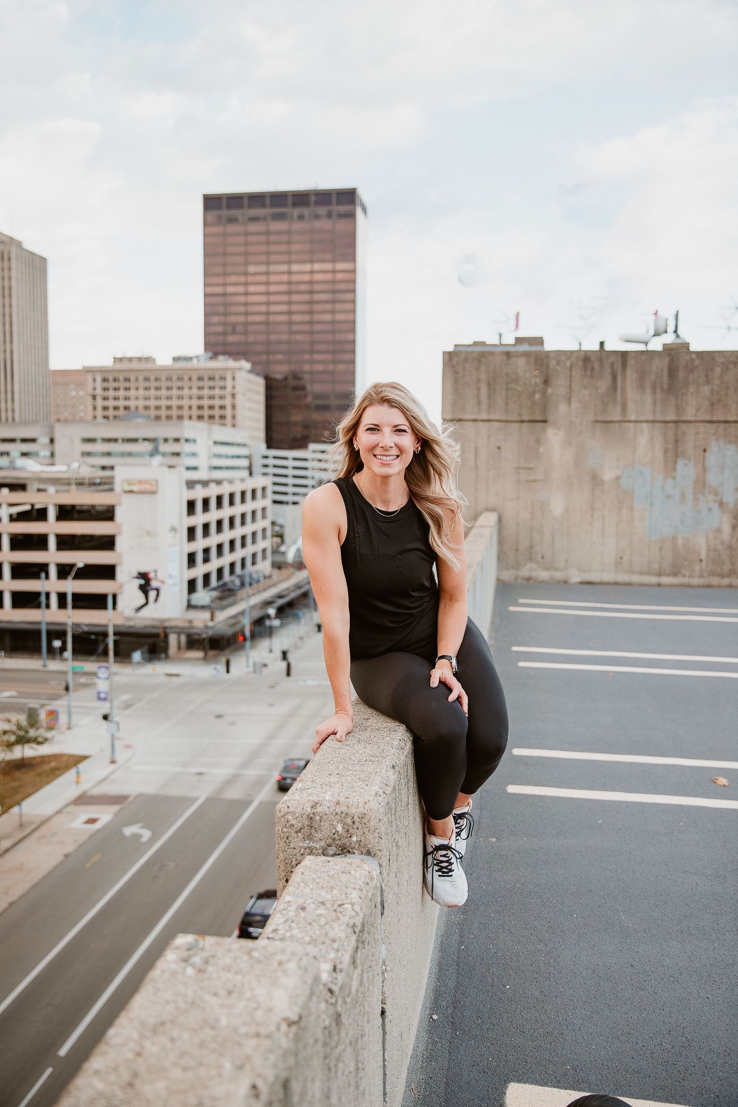 Woman in black athletic wear smiling on urban rooftop with city skyline buildings in background. Photographed by Katie Chamberlain