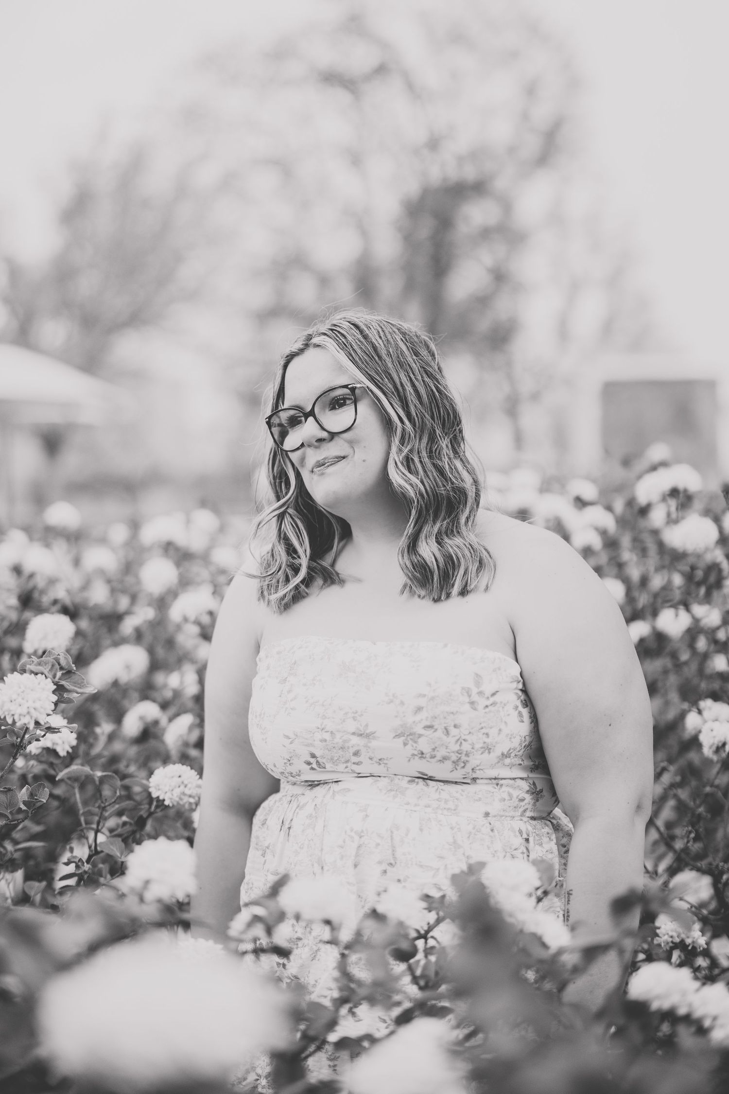 Black and white portrait of smiling woman with glasses in strapless dress among flowers. Photographed by Katie Chamberlain Photography