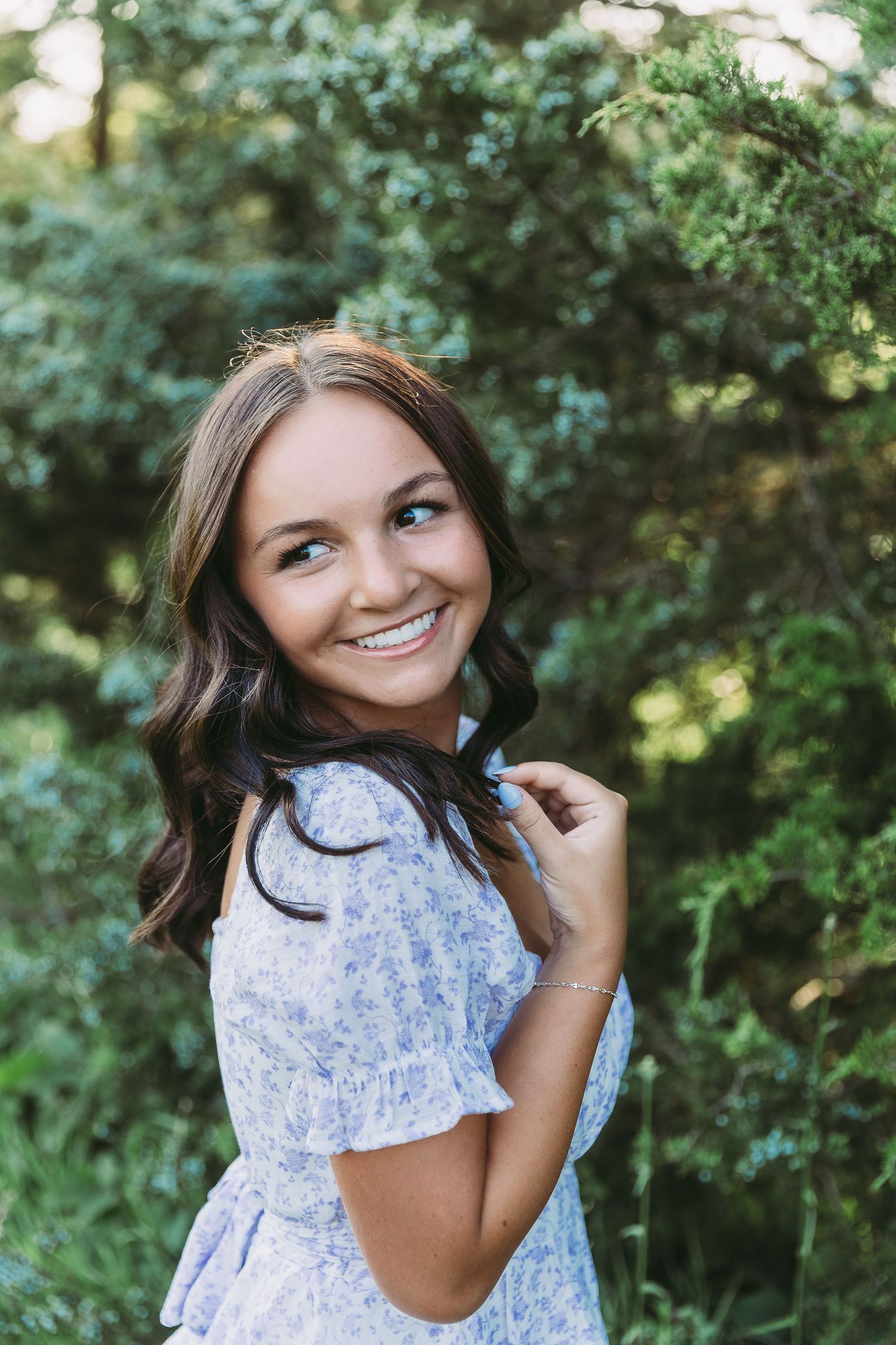 Young woman in floral blue dress smiling outdoors with lush green trees in background.