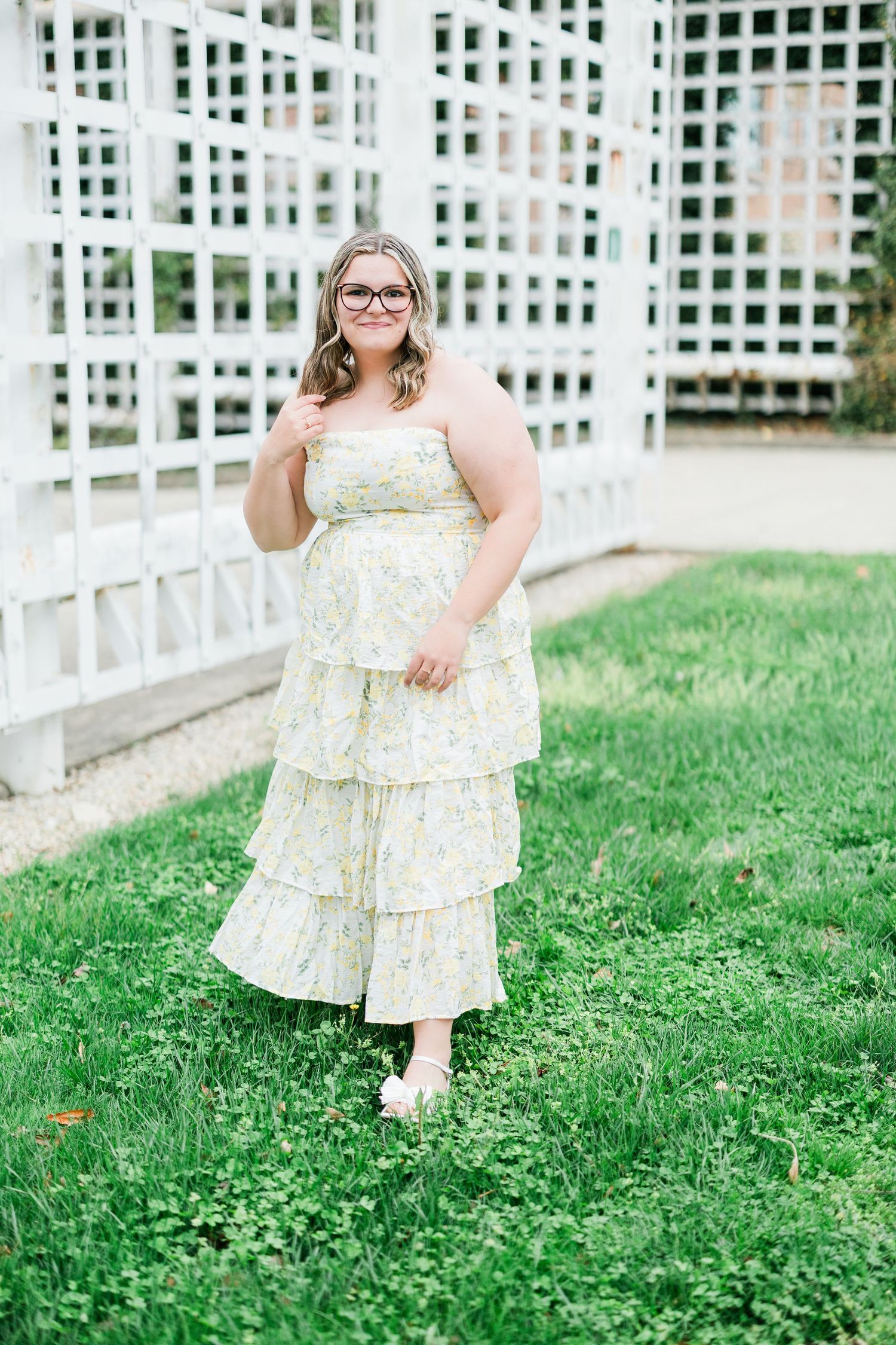 Smiling woman in yellow floral dress standing on green lawn near white garden trellis. Photographed by Katie Chamberlain Photography