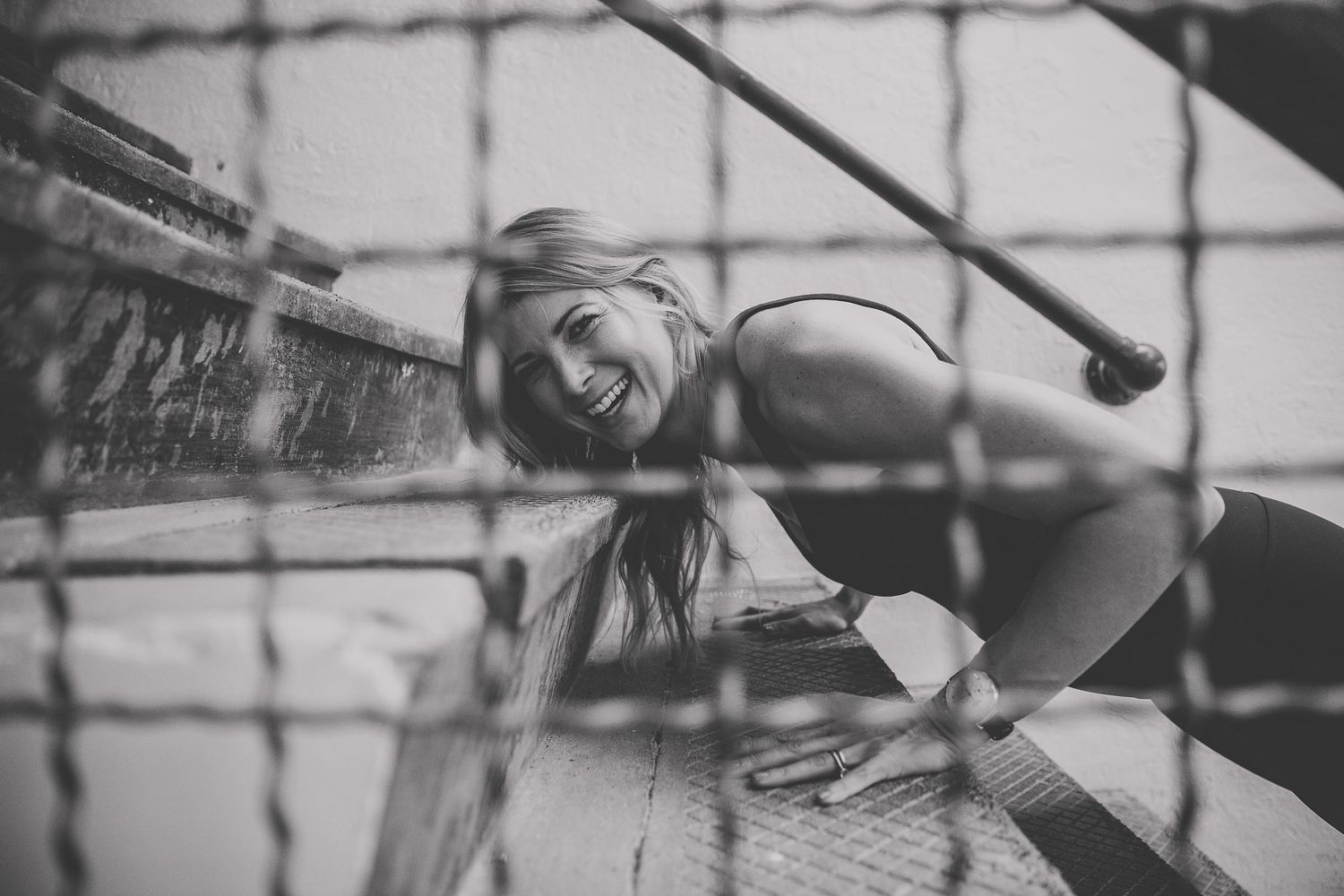 Black and white photo of a smiling blonde woman leaning on metal stairs outdoors. Photographed by Katie Chamberlain Photography
