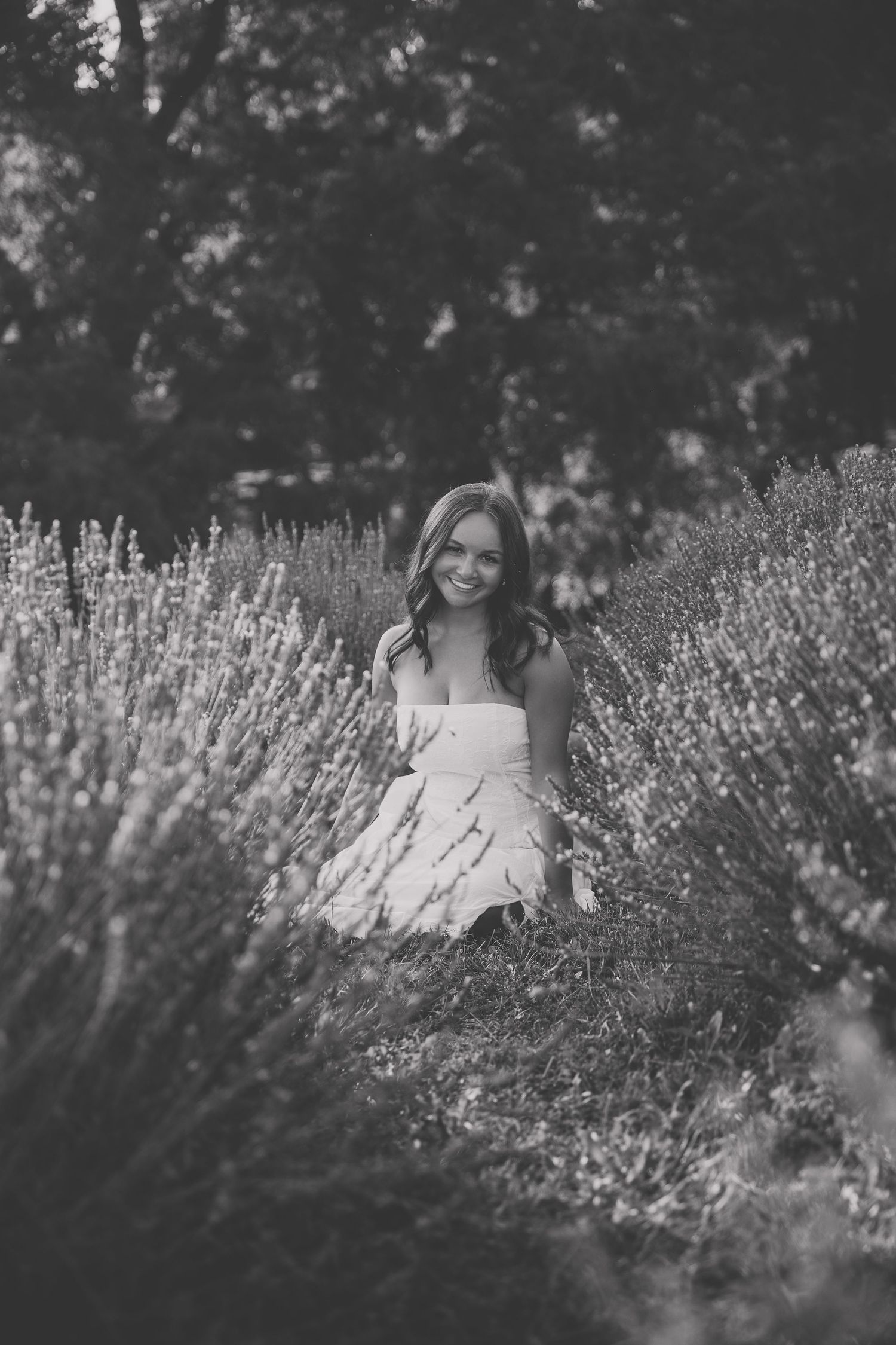 Black and white photo of a smiling young woman in a white dress sitting among lavender fields with trees in background. Katie Chamberlain