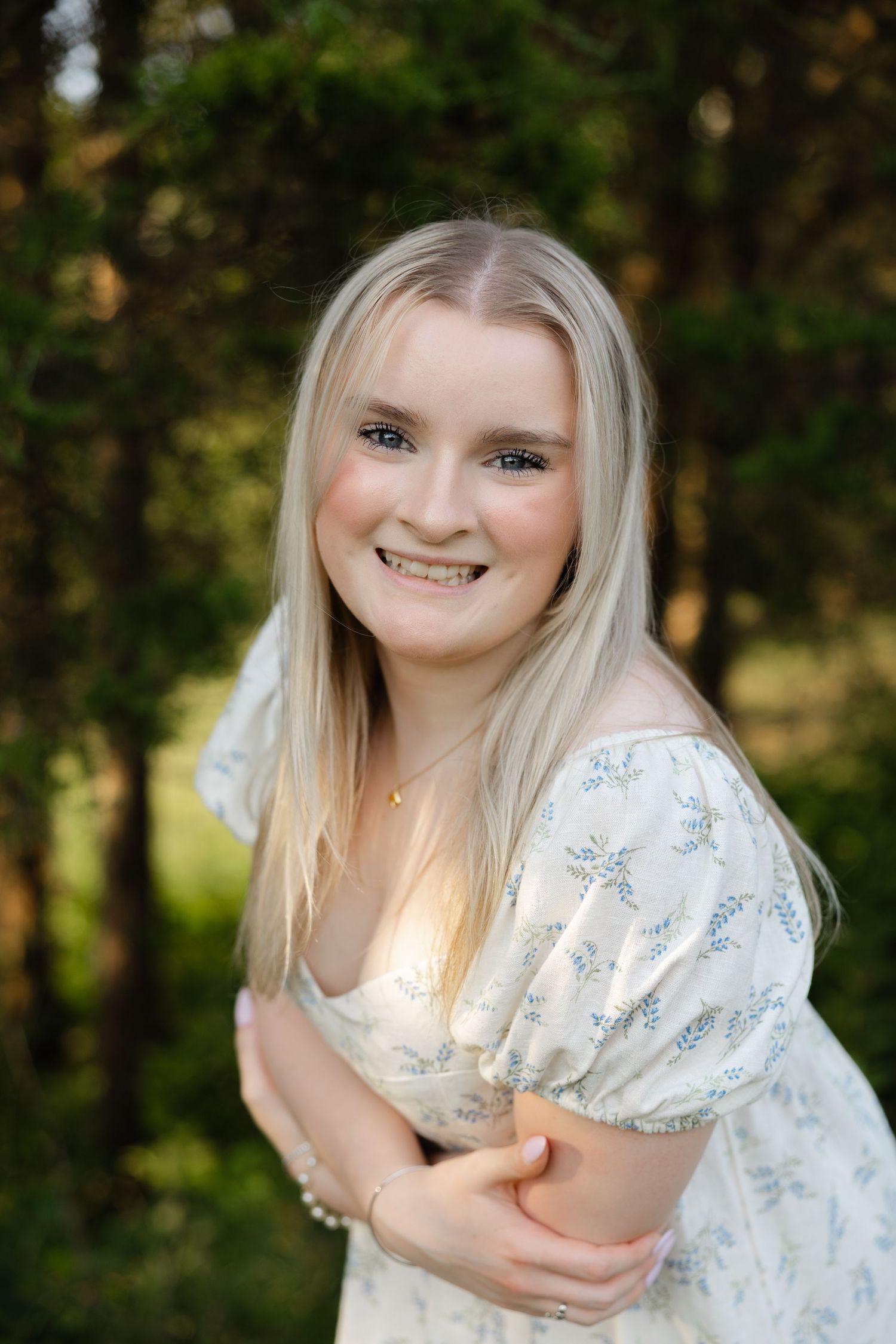 Smiling young blonde woman in a floral white dress posing outdoors among green trees.