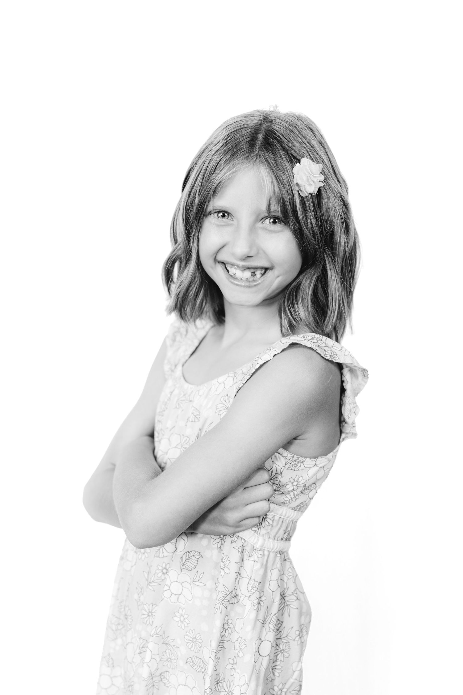 Black and white portrait of a smiling young girl with a flower in her hair, arms crossed, wearing a floral dress.