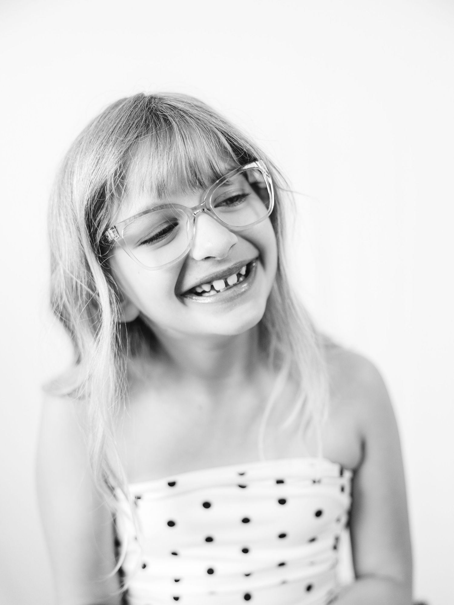 Black and white portrait of a laughing young girl with glasses and blonde bangs wearing a polka dot top.