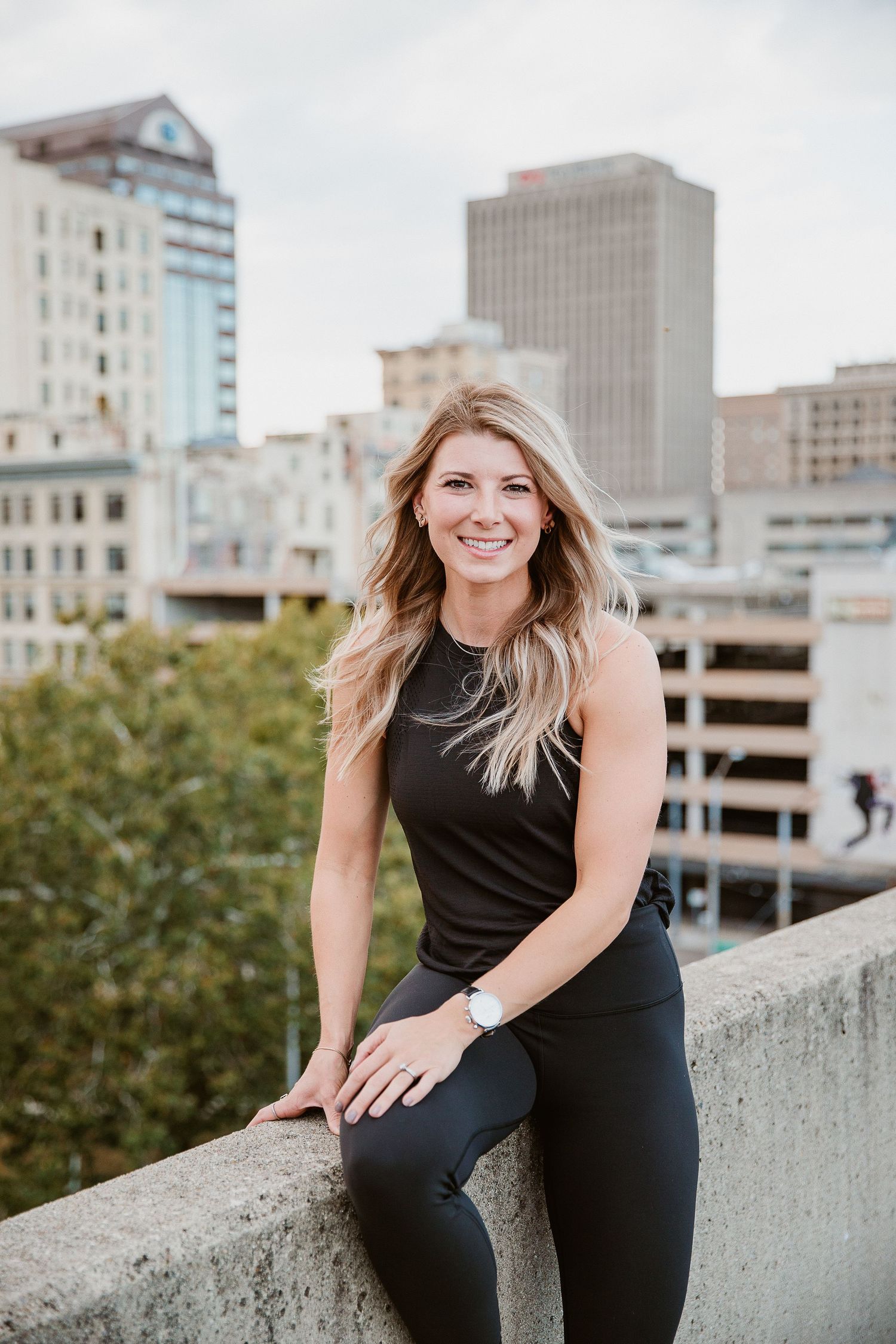Smiling blonde woman in black athletic wear posing on urban rooftop with city skyline buildings in background. Photo by Katie Chamberlain