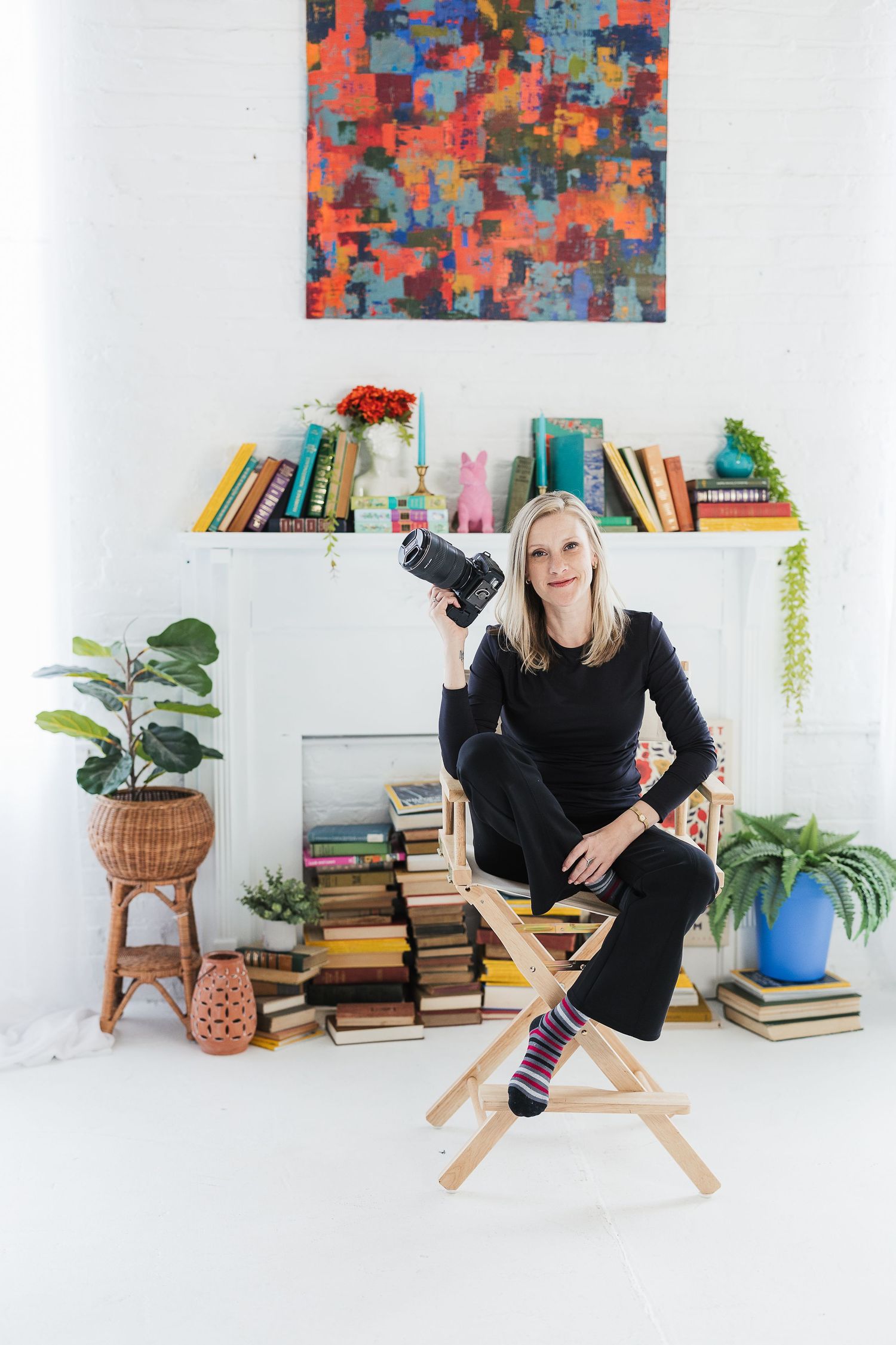 Woman in black outfit holds camera, sits on stool surrounded by books, plants, and colorful art in bright white room. Katie Chamberlain