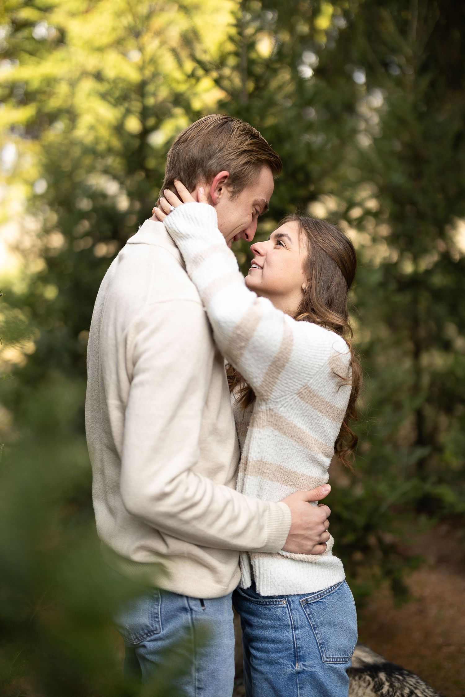 A couple in cream sweaters and jeans embrace and smile at each other in a lush green forest setting. Photographed by Katie Chamberlain Photo