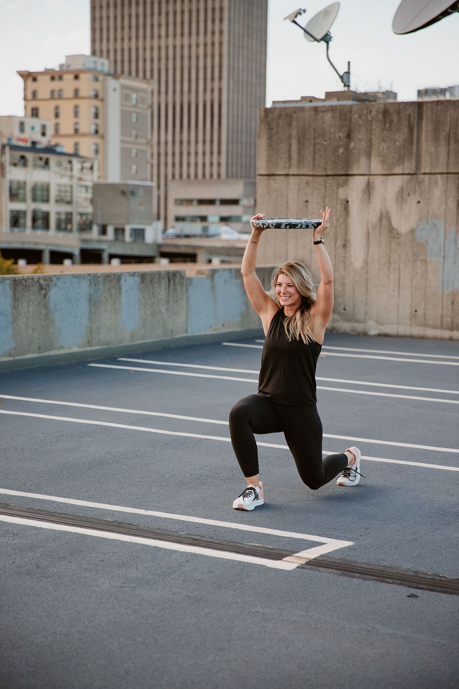 Woman in black workout attire doing a lunge on a rooftop parking deck with city buildings in the background. Photo by Katie Chamberlain