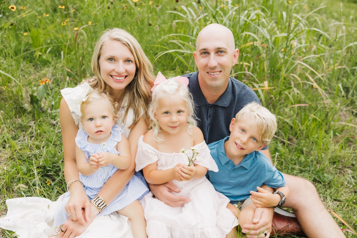 Happy family of five smiling outdoors in a green meadow, parents with three young children on a sunny day.