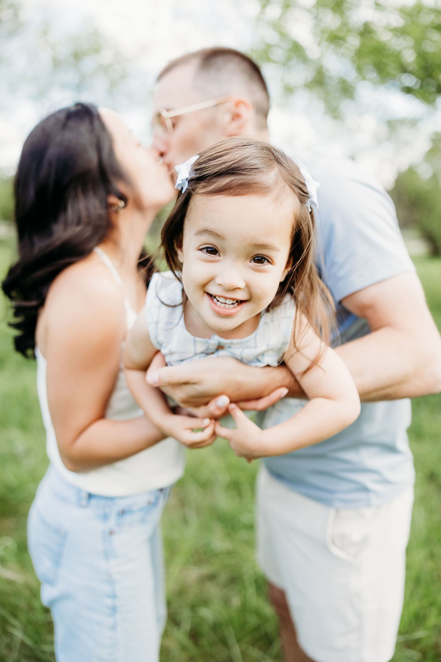 Happy toddler girl smiling while parents kiss her cheek outdoors in a green park setting.