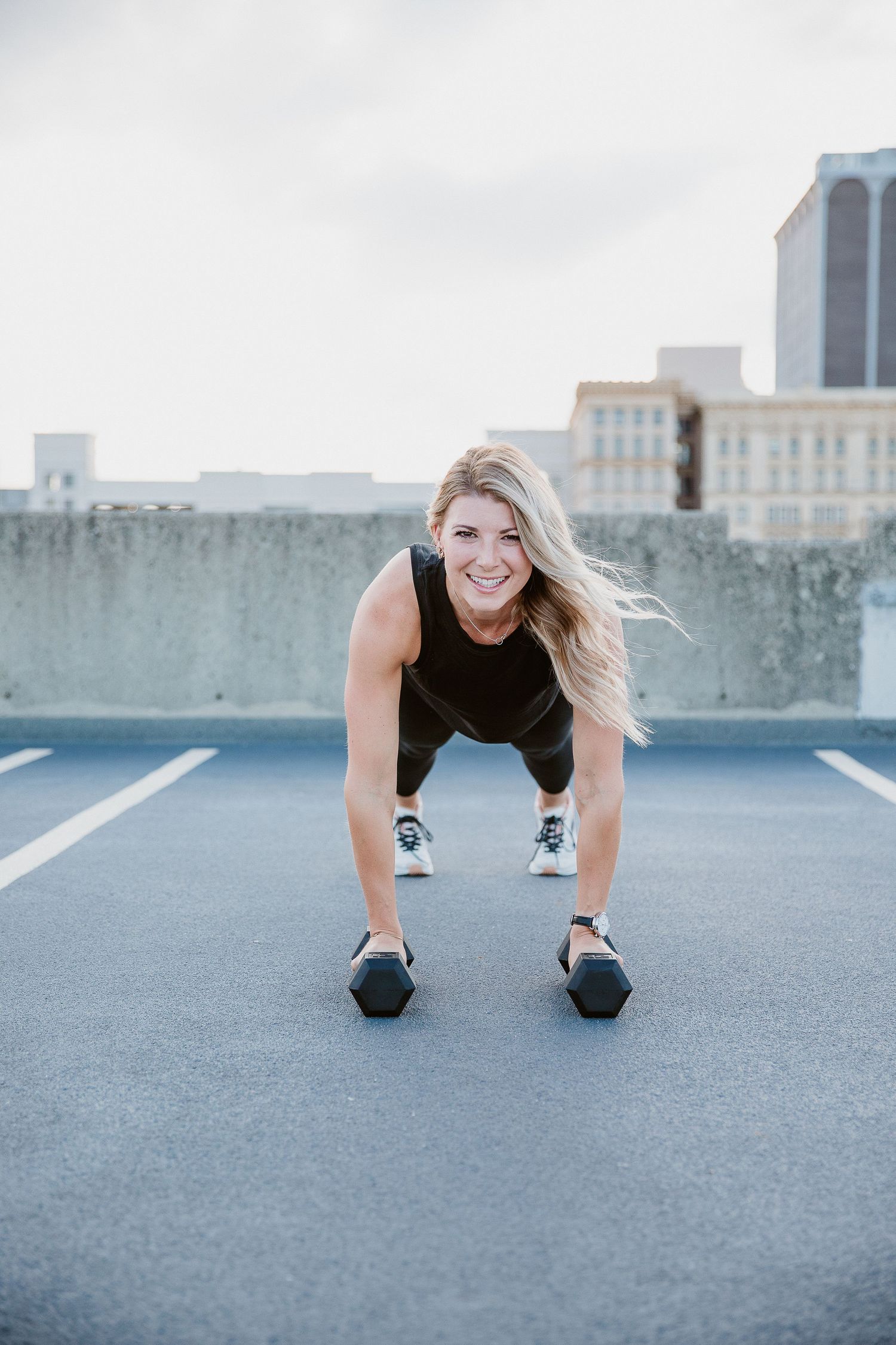 Smiling blonde woman in black athletic wear doing dumbbell deadlifts on rooftop with city skyline background. Photo by Katie Chamberlain