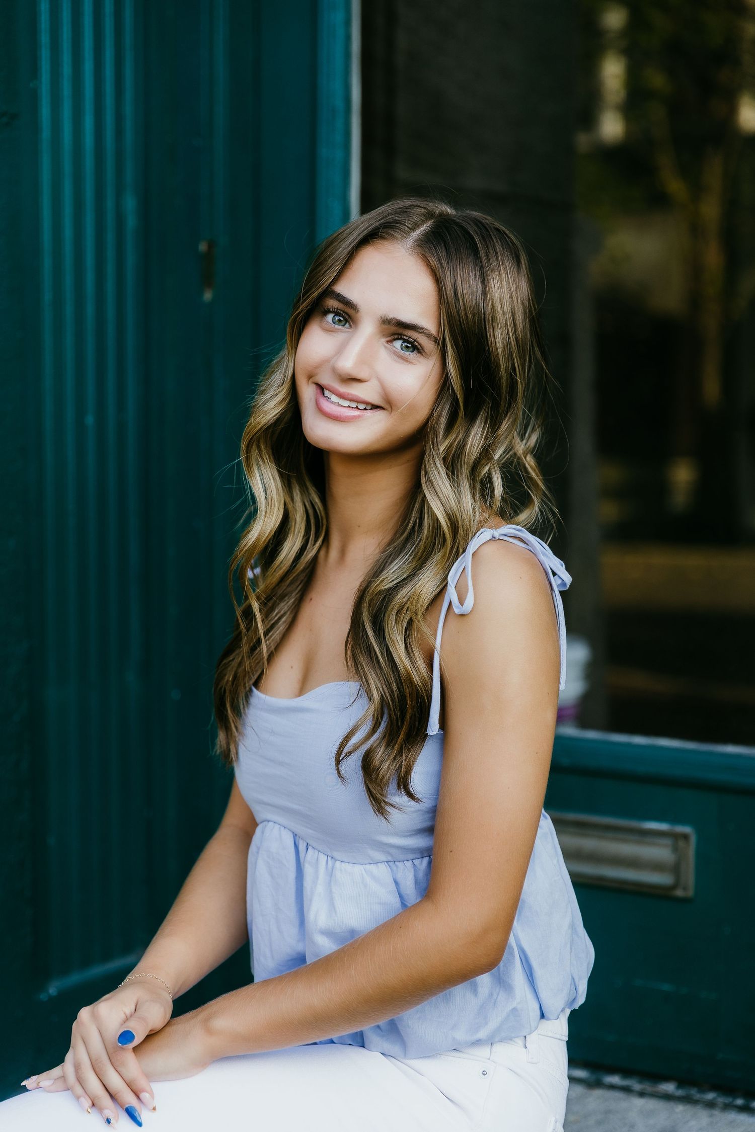 Young woman with wavy brown hair smiles in a light blue top and white pants, seated near a teal door. Katie Chamberlain Photography