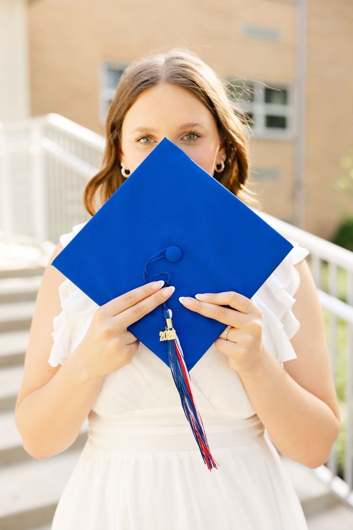 Young woman in white dress holding blue graduation cap in front of her face outdoors.