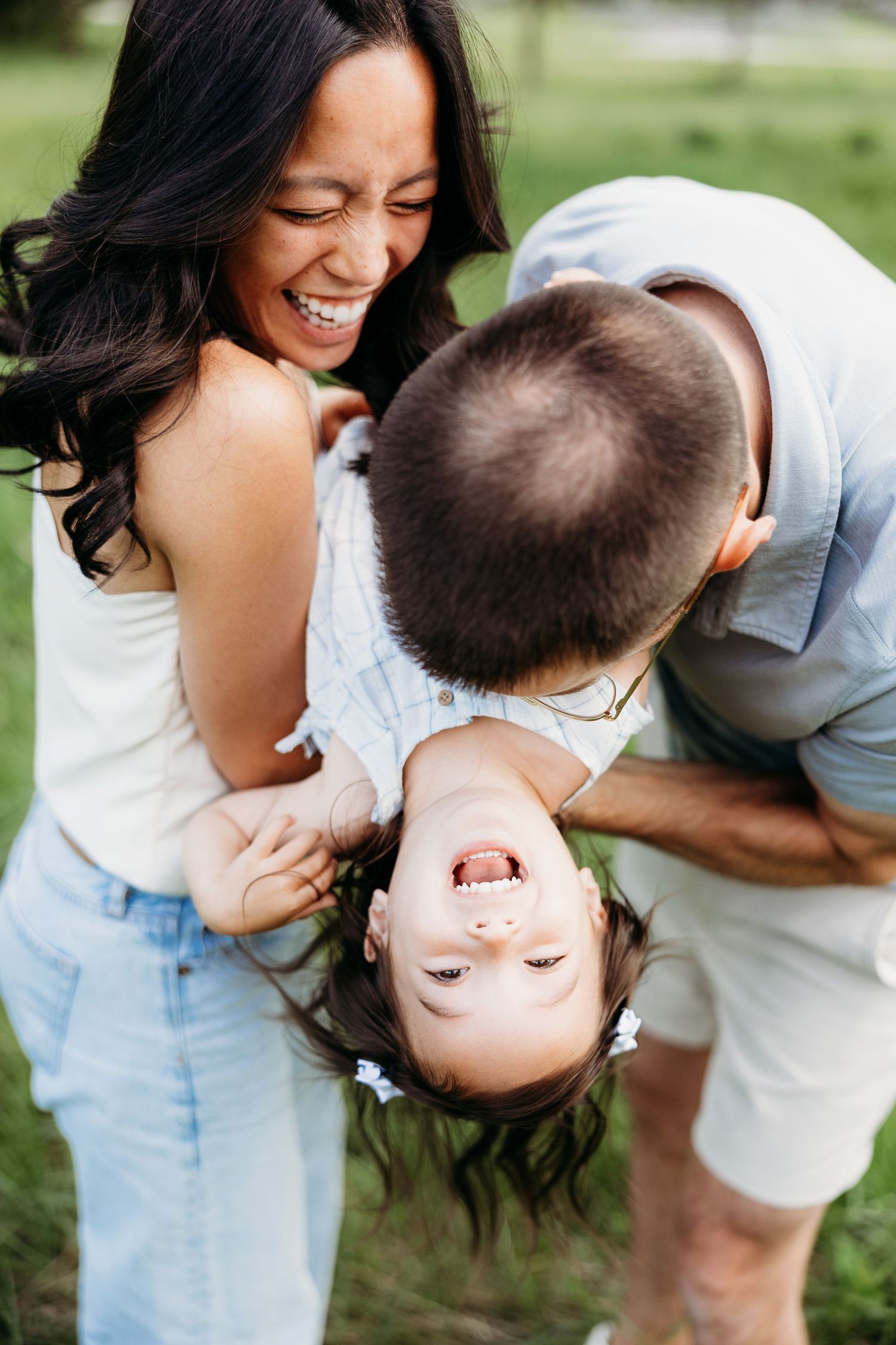 Laughing parents playfully flip their giggling toddler upside down in a sunny green outdoor setting.