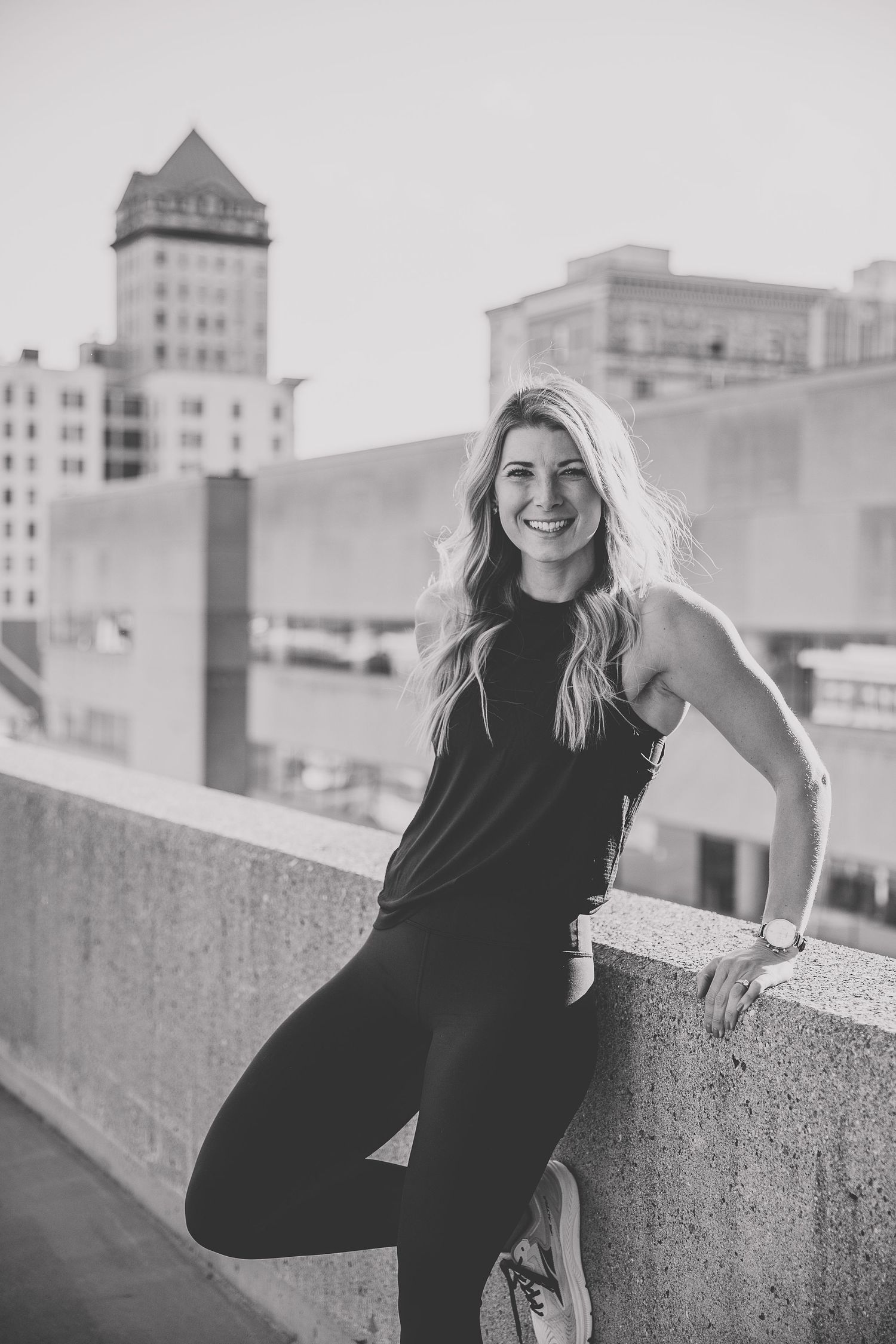 Smiling blonde woman in black athletic wear leaning against a concrete wall urban buildings in the background. Photo by Katie Chamberlain