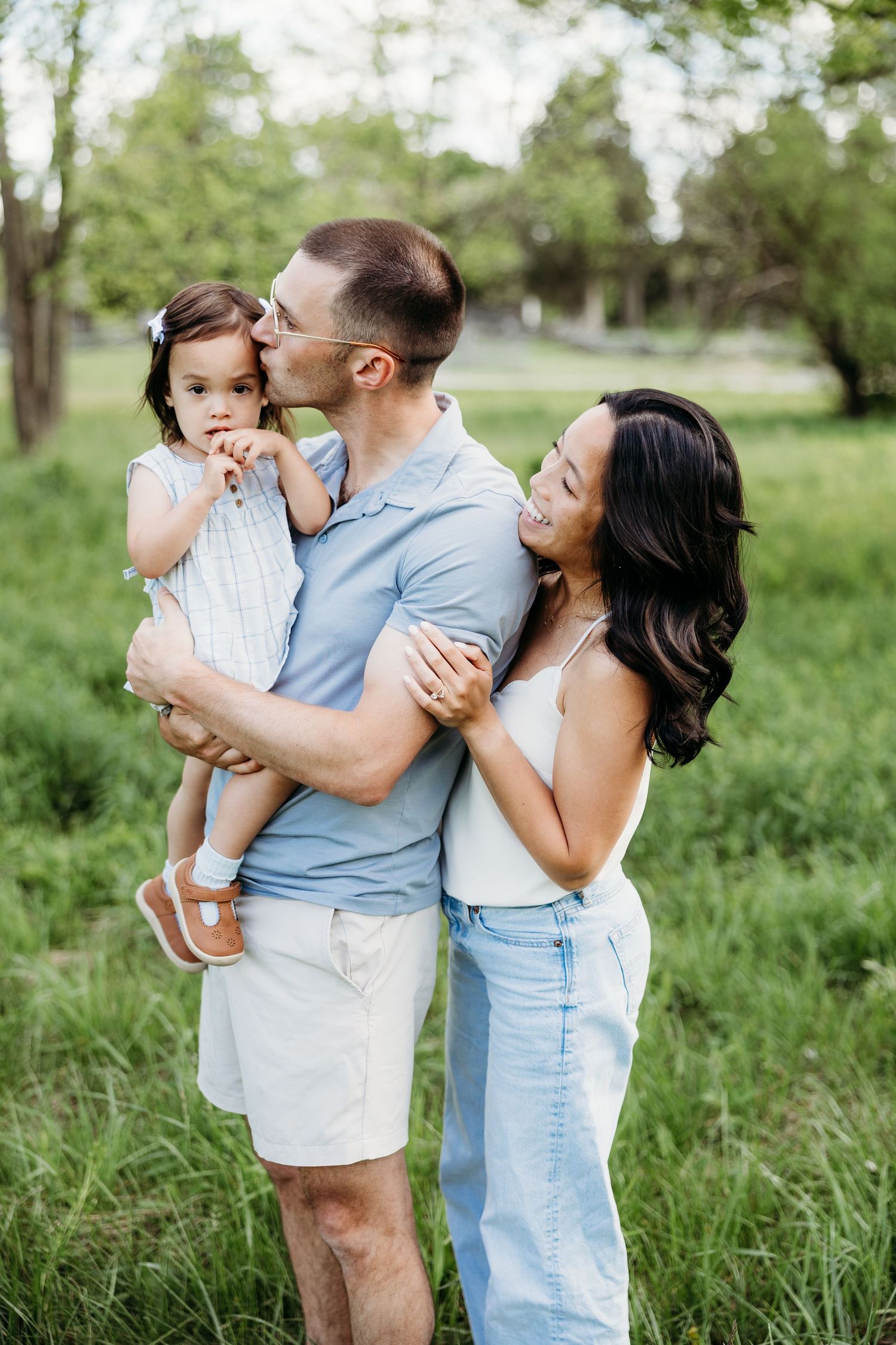 A happy family poses outdoors, father kissing toddler daughter while mother smiles in a lush green park.