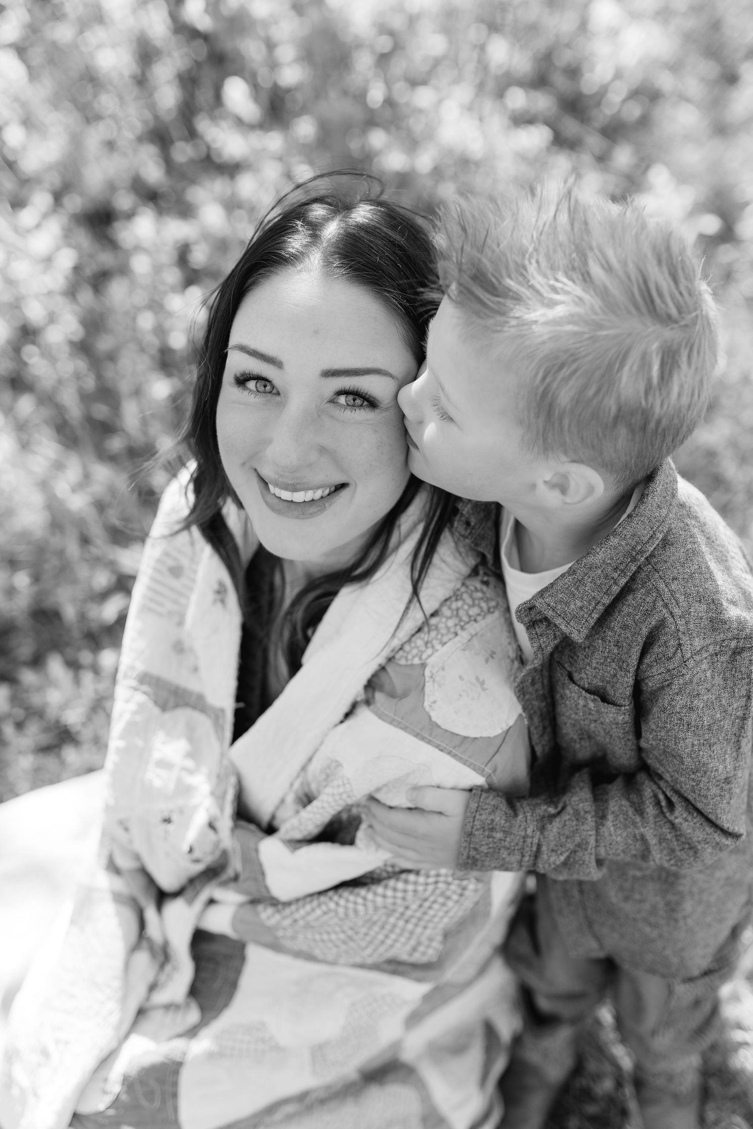 Black and white photo of a smiling woman wrapped in a quilt as a young blonde boy kisses her cheek outdoors.