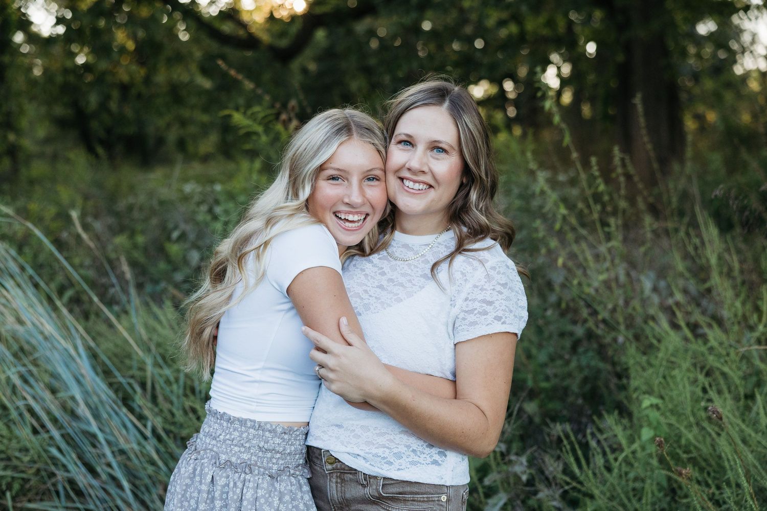 Two smiling young women in white tops hugging outdoors in a lush green field at golden hour.