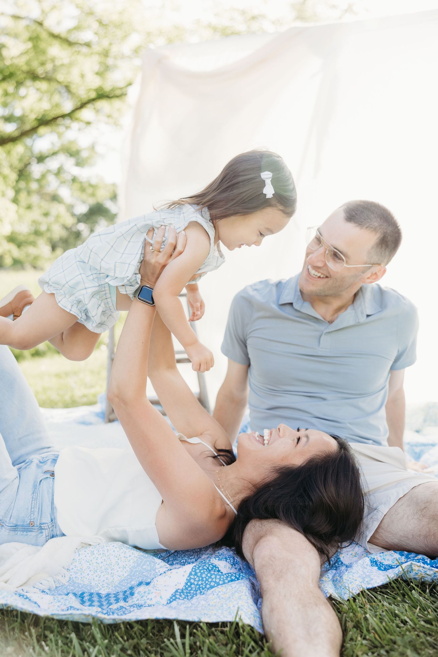 Family enjoying a sunny picnic on a blanket, with parents playfully lifting their young daughter outdoors.