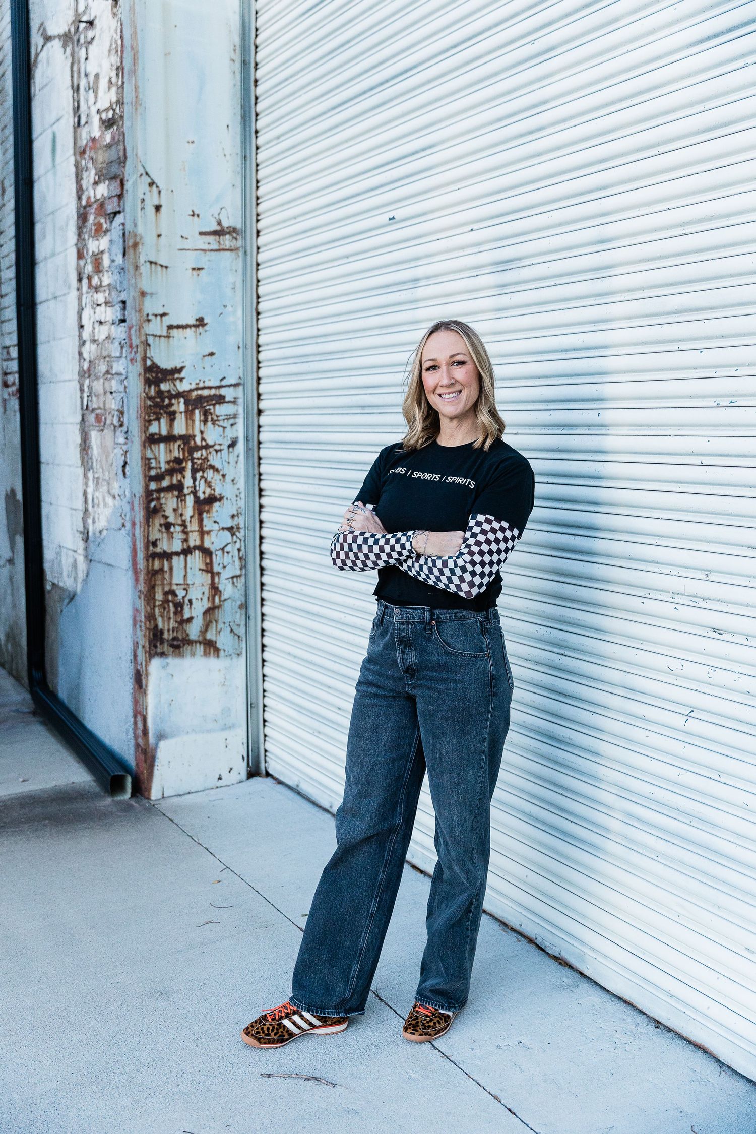 Smiling blonde woman in black Subs Sports Spirits t-shirt with checkered sleeves, arms crossed, posing in urban alley. Katie Chamberlain