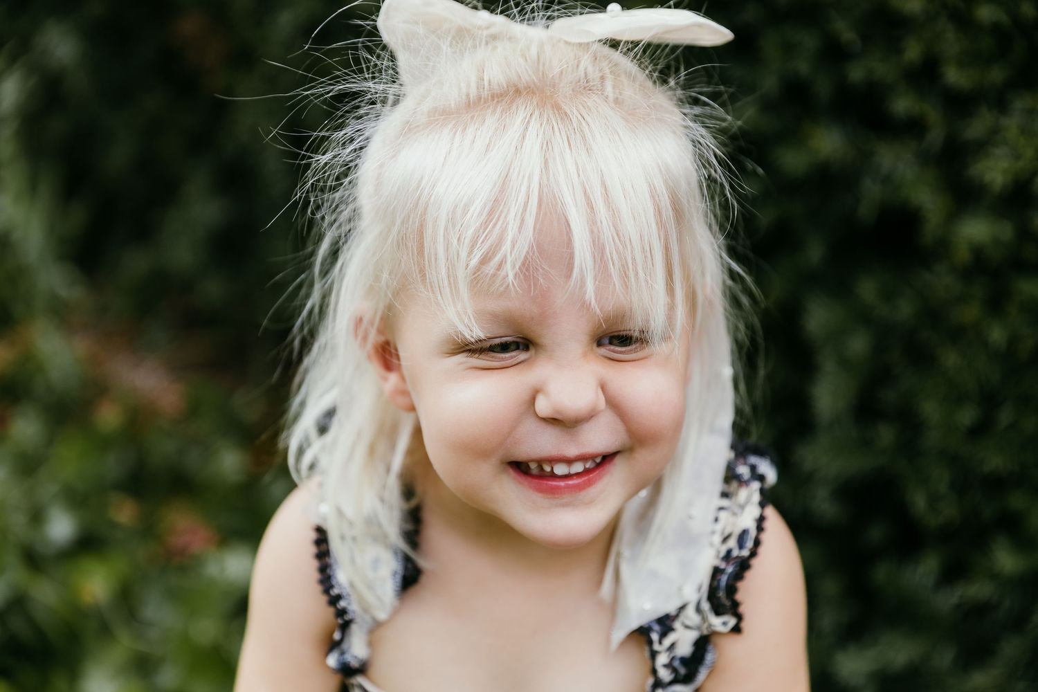 Smiling blonde toddler girl with hair bow outdoors, wearing floral top, green foliage background. Photographed by Katie Chamberlain