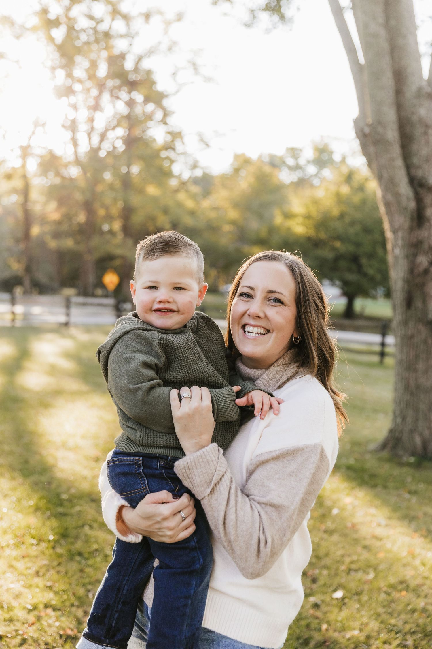 A smiling woman holds a happy toddler boy outdoors in a sunny park during autumn. Katie Chamberlain Photography