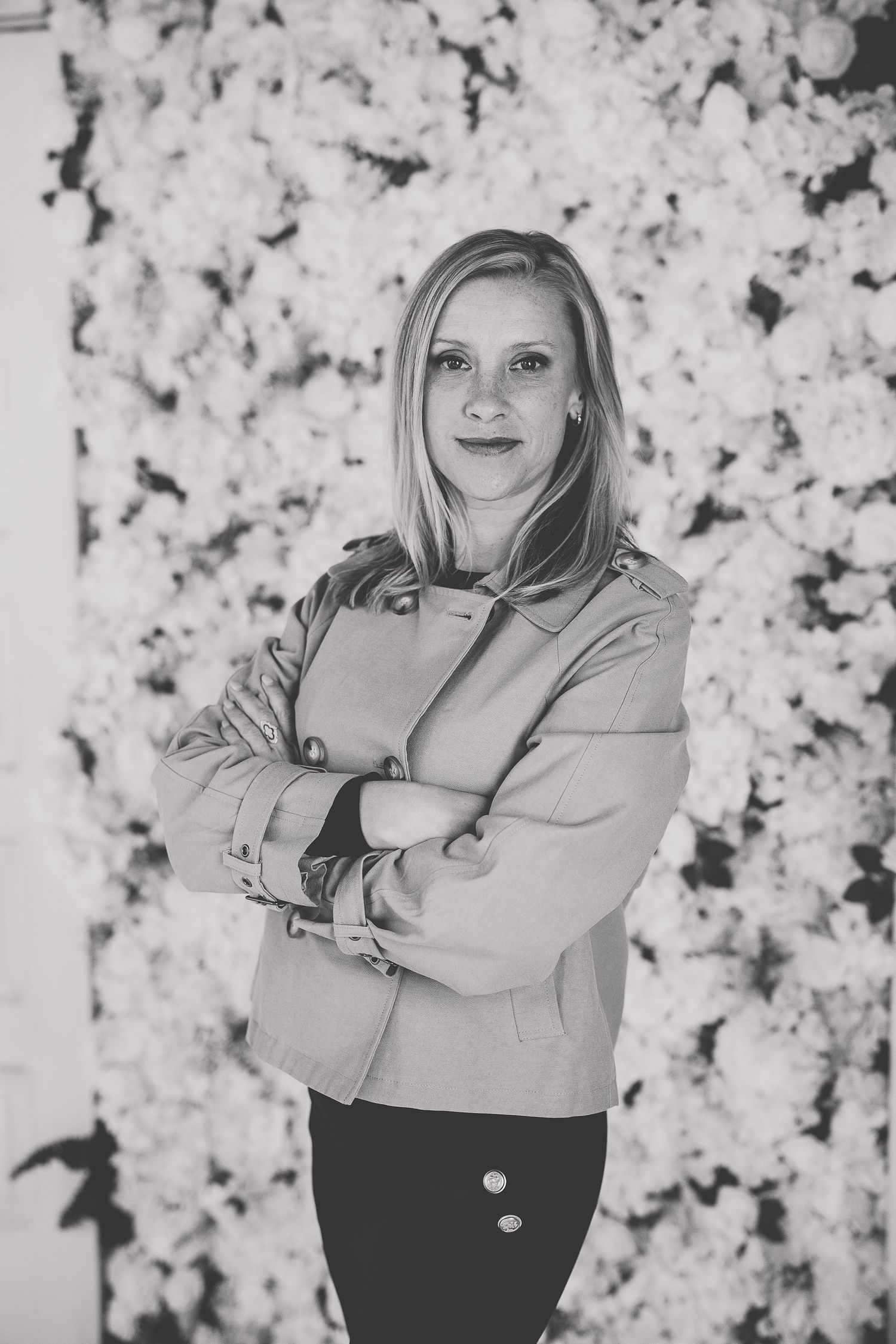Black and white portrait of a blonde woman with arms crossed standing in front of a white floral wall backdrop. Katie Chamberlain Photograph