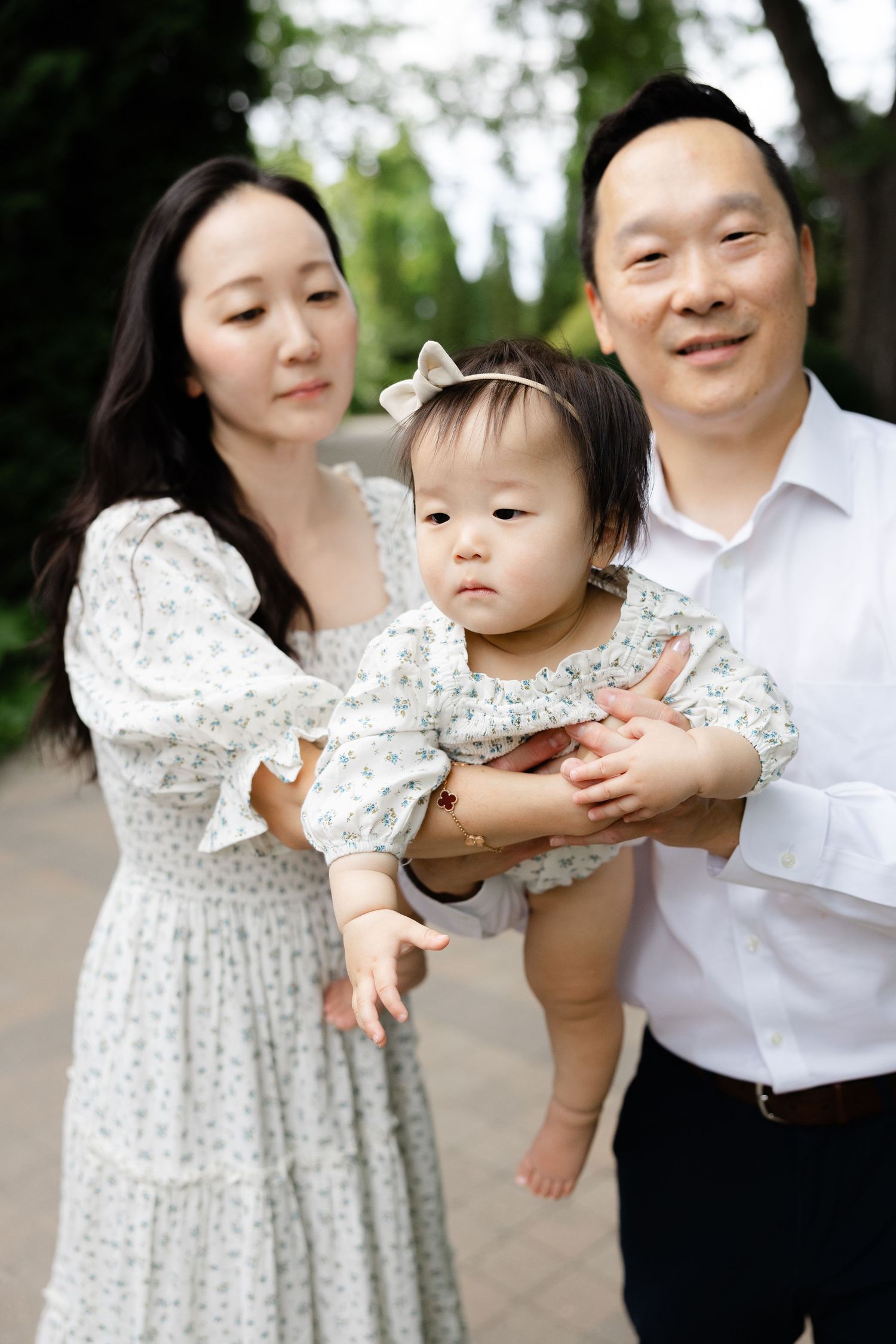 Asian family portrait with mother in floral dress and father in white shirt holding baby girl outdoors.