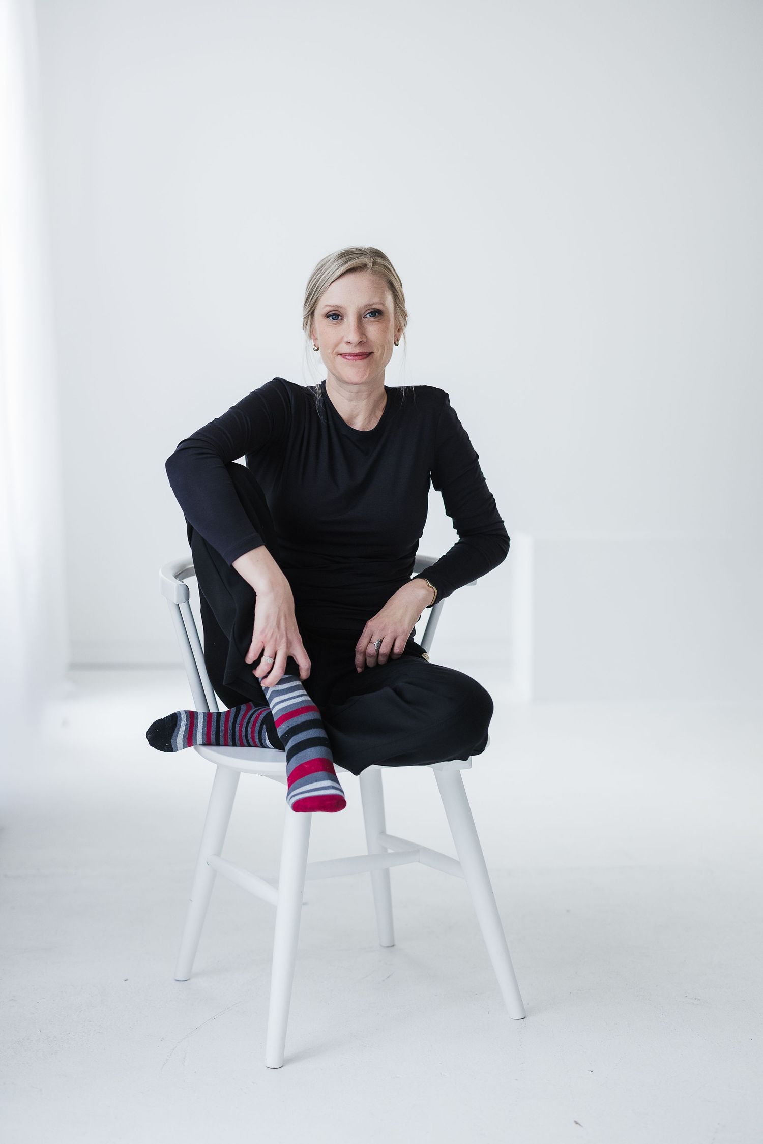 Woman in black outfit with colorful striped socks sitting cross-legged on white chair against white background. Katie Chamberlain Photograph