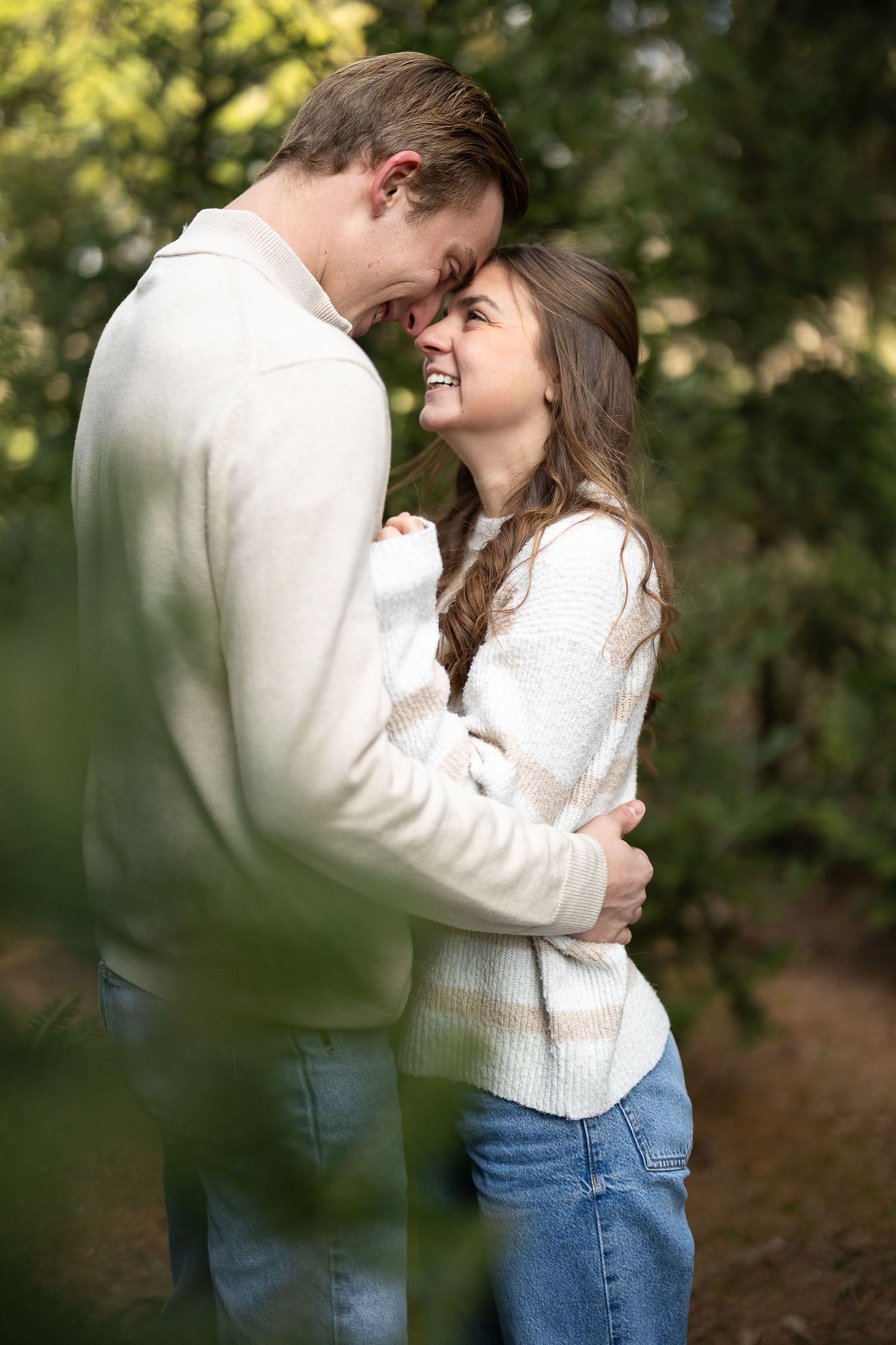 Couple embracing and smiling at each other outdoors, wearing cozy sweaters and jeans among green trees. Photographed by Katie Chamberlain