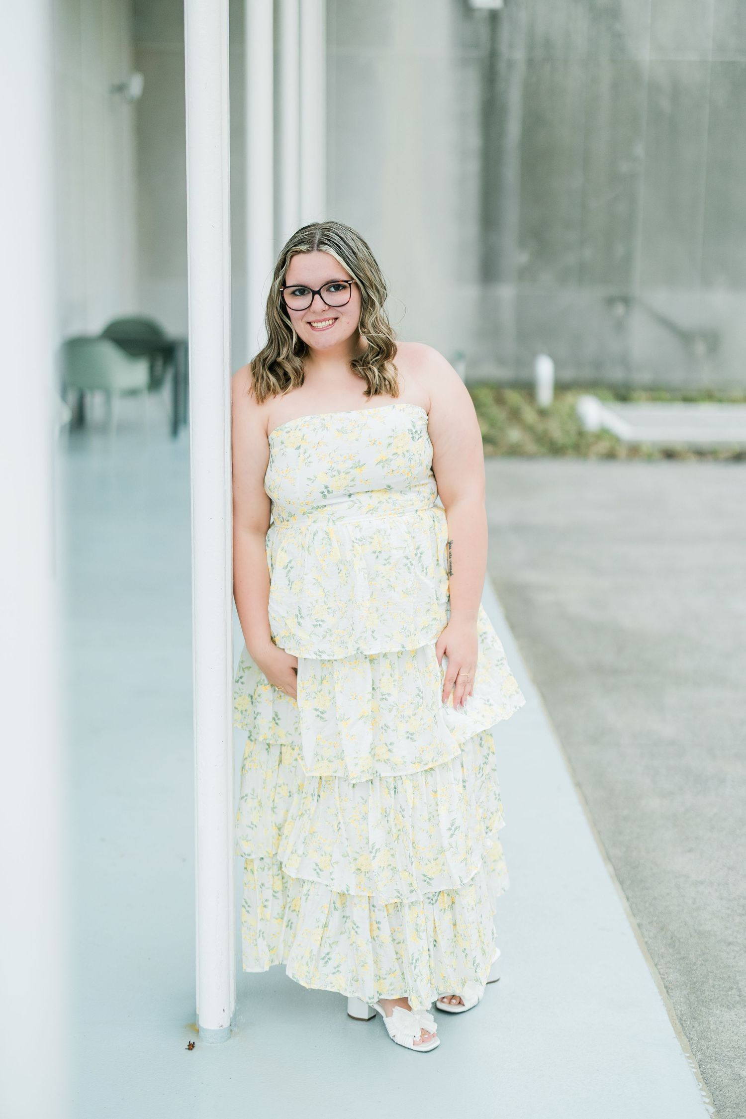 Young woman in yellow floral tiered maxi dress leaning against white column outdoors. Photographed by Katie Chamberlain Photography